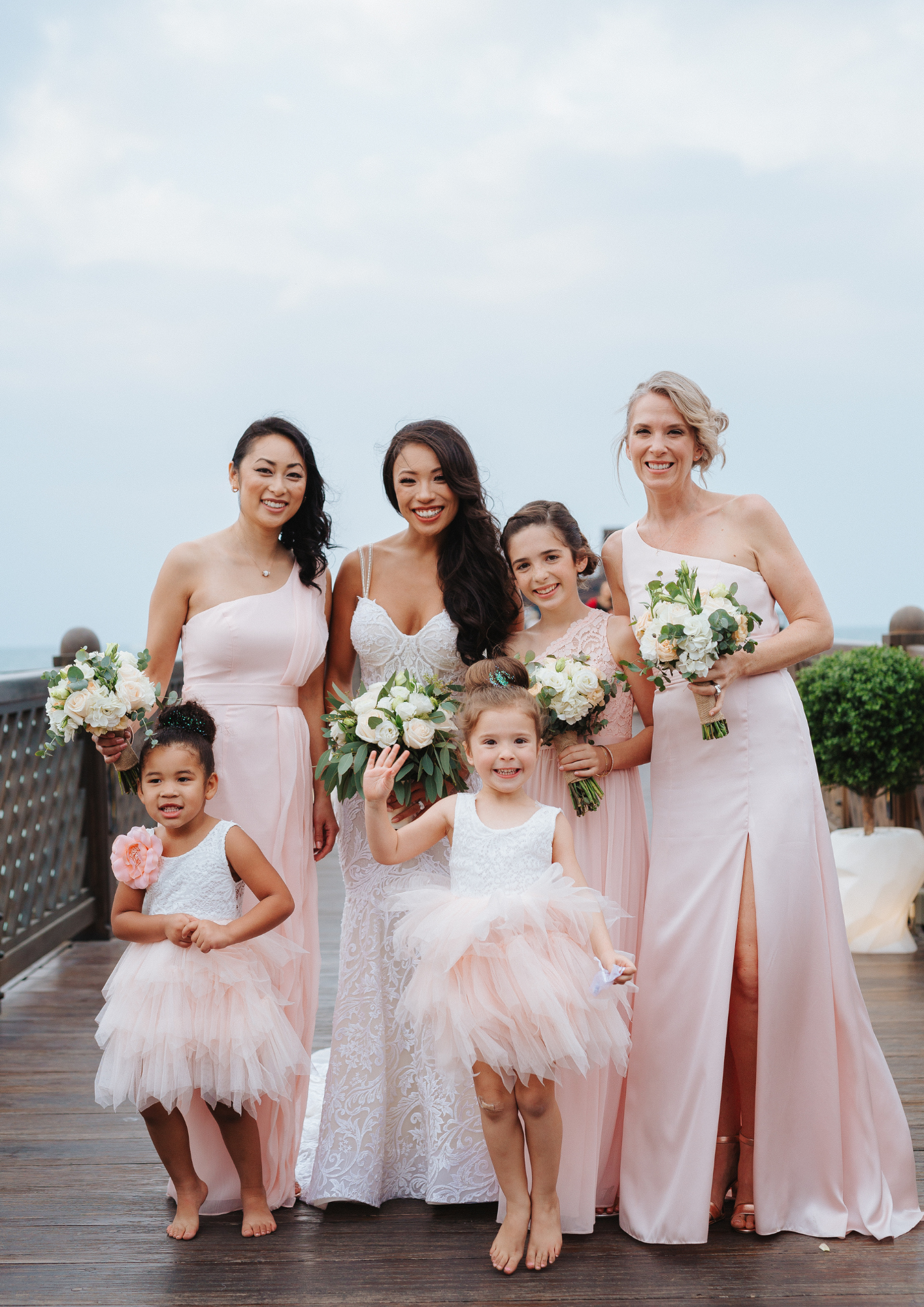 Group of women and children celebrating a wedding outdoors, dressed in pink and white dresses, holding bouquets of flowers.
