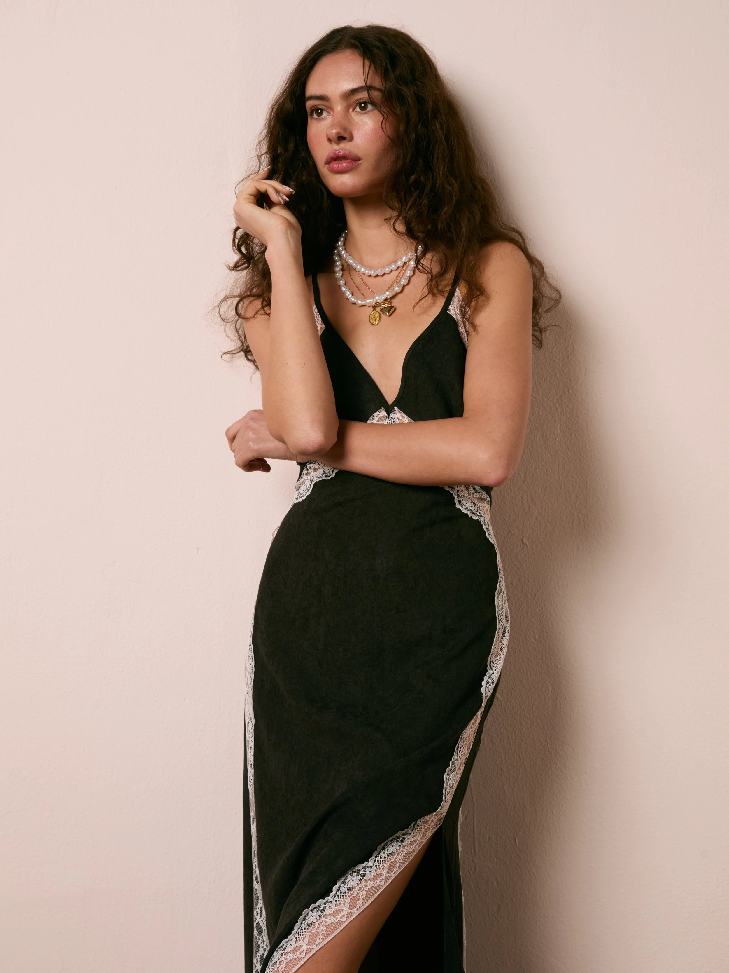Woman with curly brown hair wearing a black dress with lace details, layered necklaces, and standing against a plain wall.