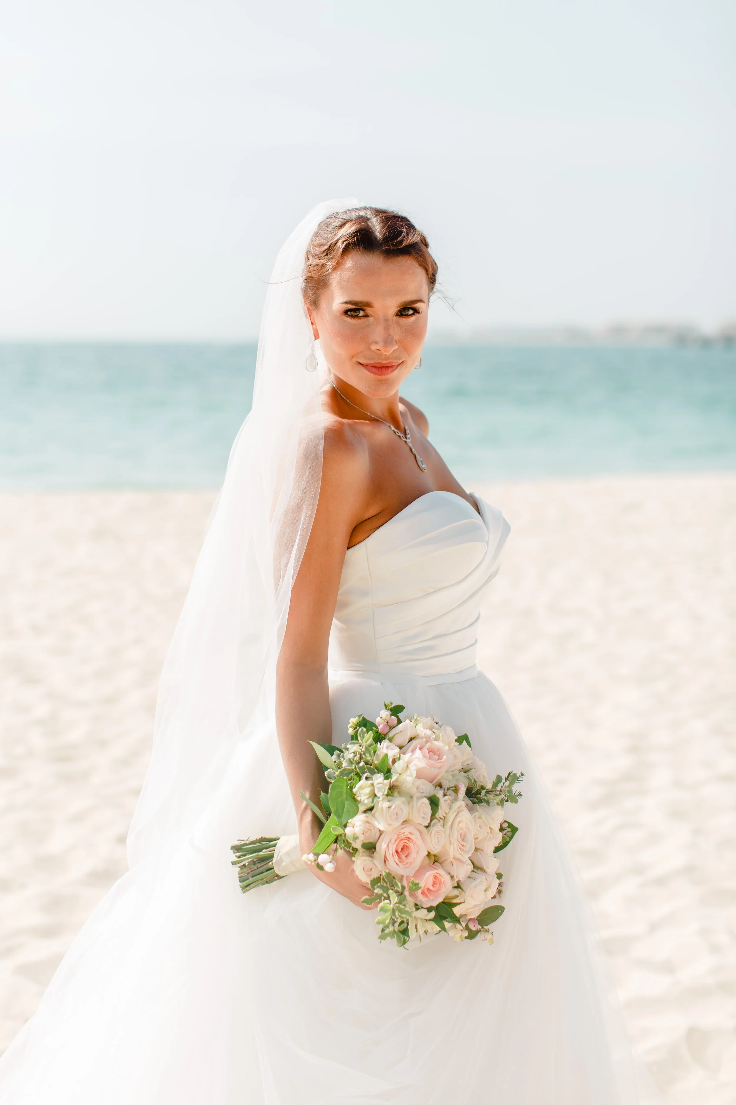 Bride in a white wedding gown holding a bouquet of pink and white roses on a beach.