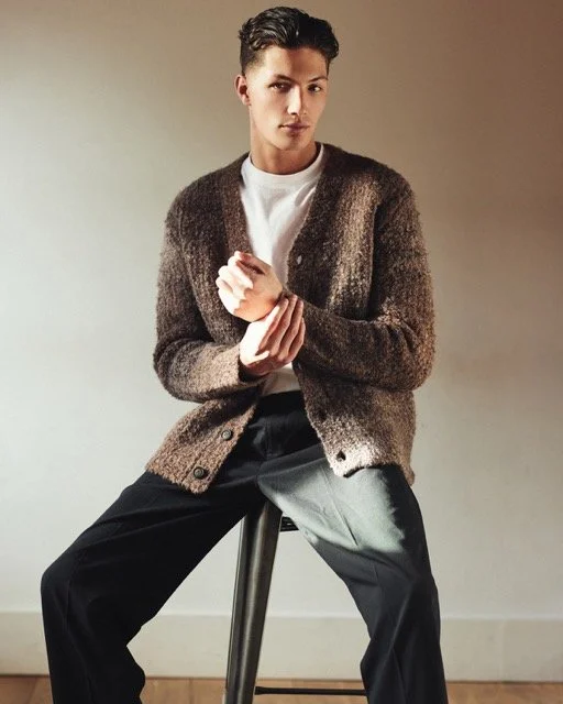 A young man sitting on a stool in a studio, wearing a white shirt, brown cardigan, and black pants, looking directly at the camera.
