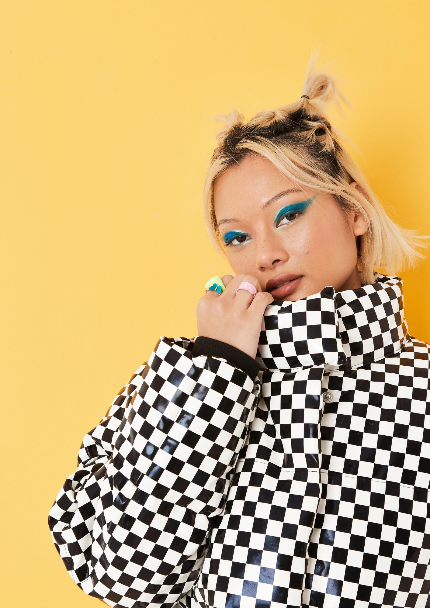 A woman with blonde hair styled in small buns, wearing a black and white checkered puffer jacket, poses with her hand near her face against a yellow background with vibrant blue eye makeup.