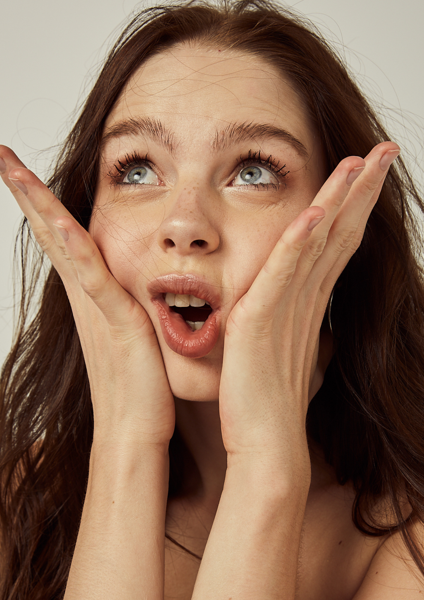 A woman with brown hair and blue eyes holding her face with her hands, expressing surprise.