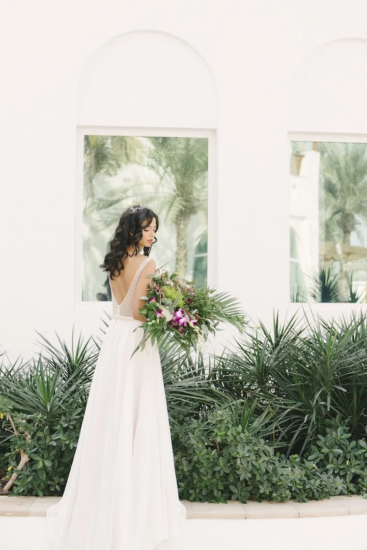 Woman in a white dress holding a large bouquet standing in front of a white building with large windows and green plants.