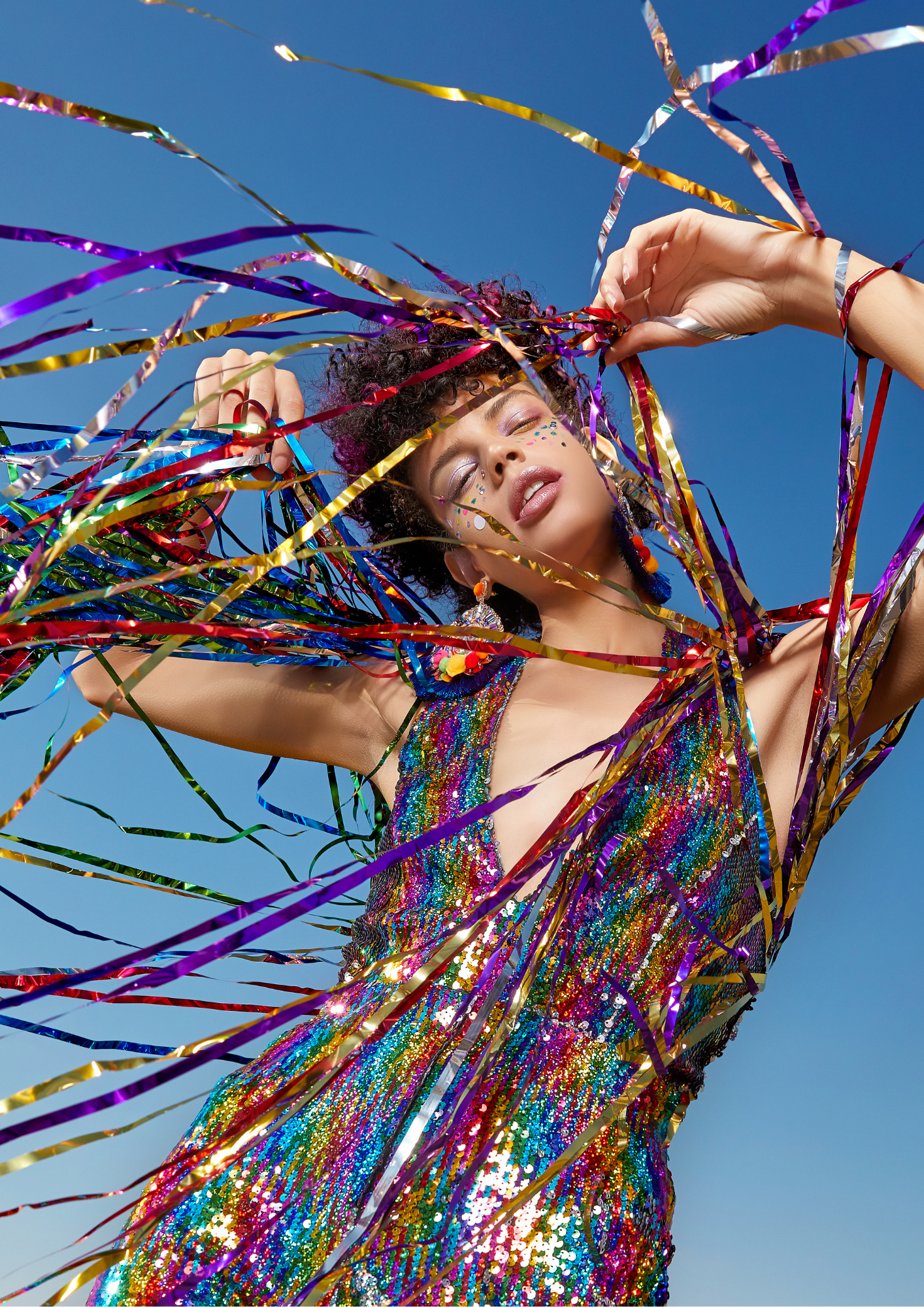 Woman with closed eyes and colorful makeup, wearing a sequined dress, surrounded by rainbow-colored streamers against a clear blue sky.