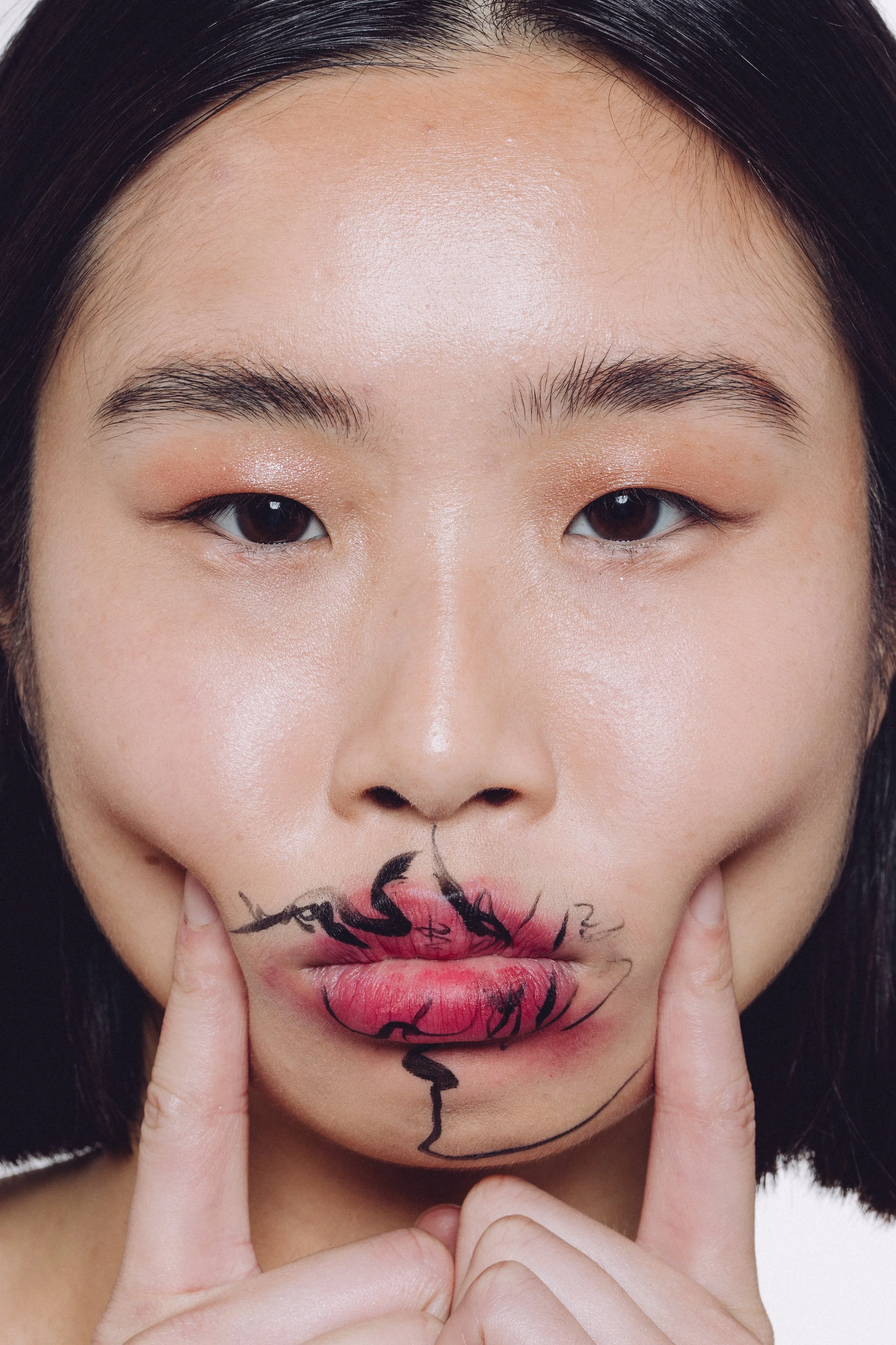 Close-up of a woman with dark hair and makeup, holding her lips with her index fingers. Her lips have artistic black ink drawings and pink coloring around them.