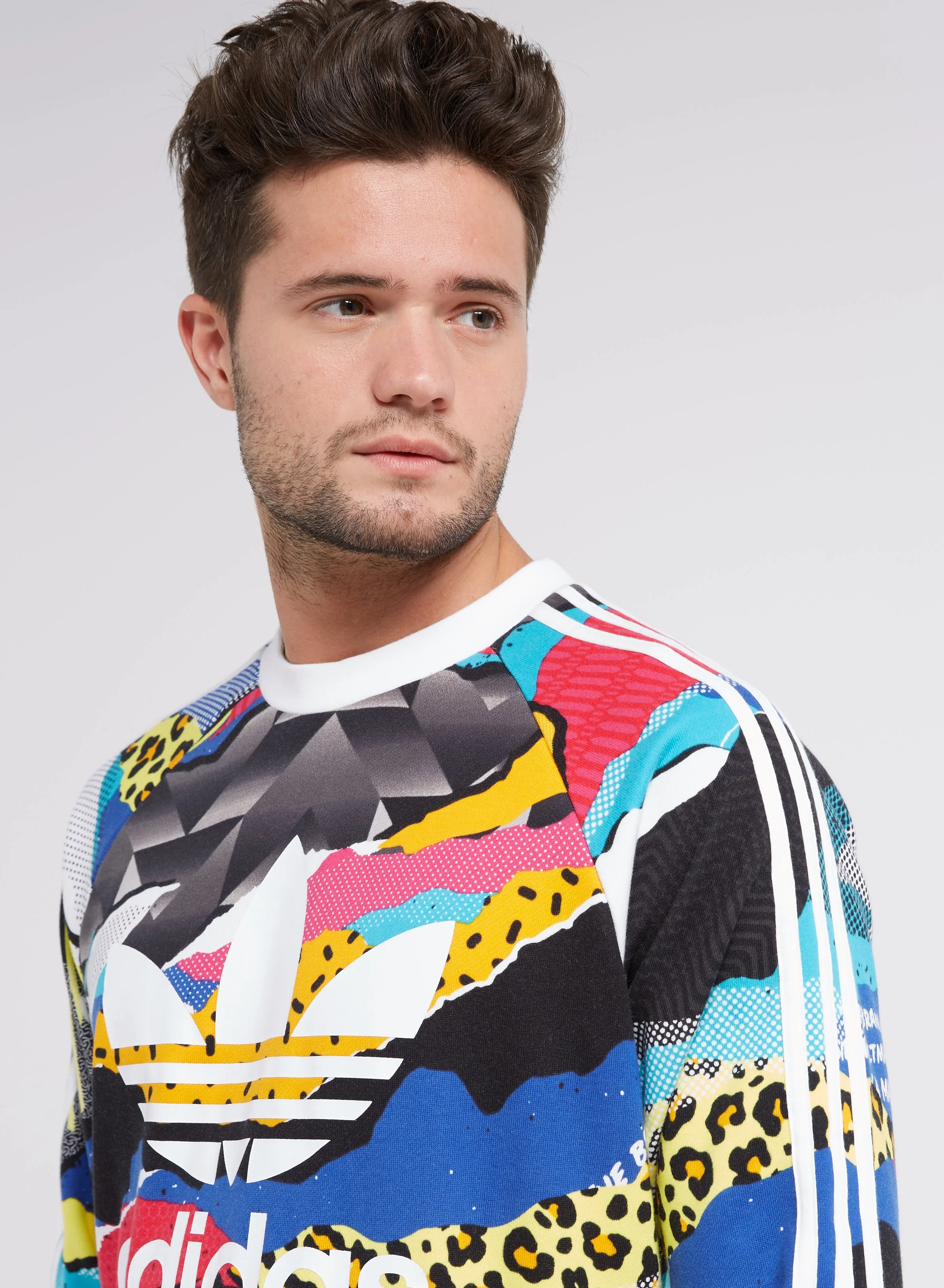 A young man with brown hair and facial stubble wearing a colorful graphic Adidas sweatshirt, looking to the side against a plain gray background.
