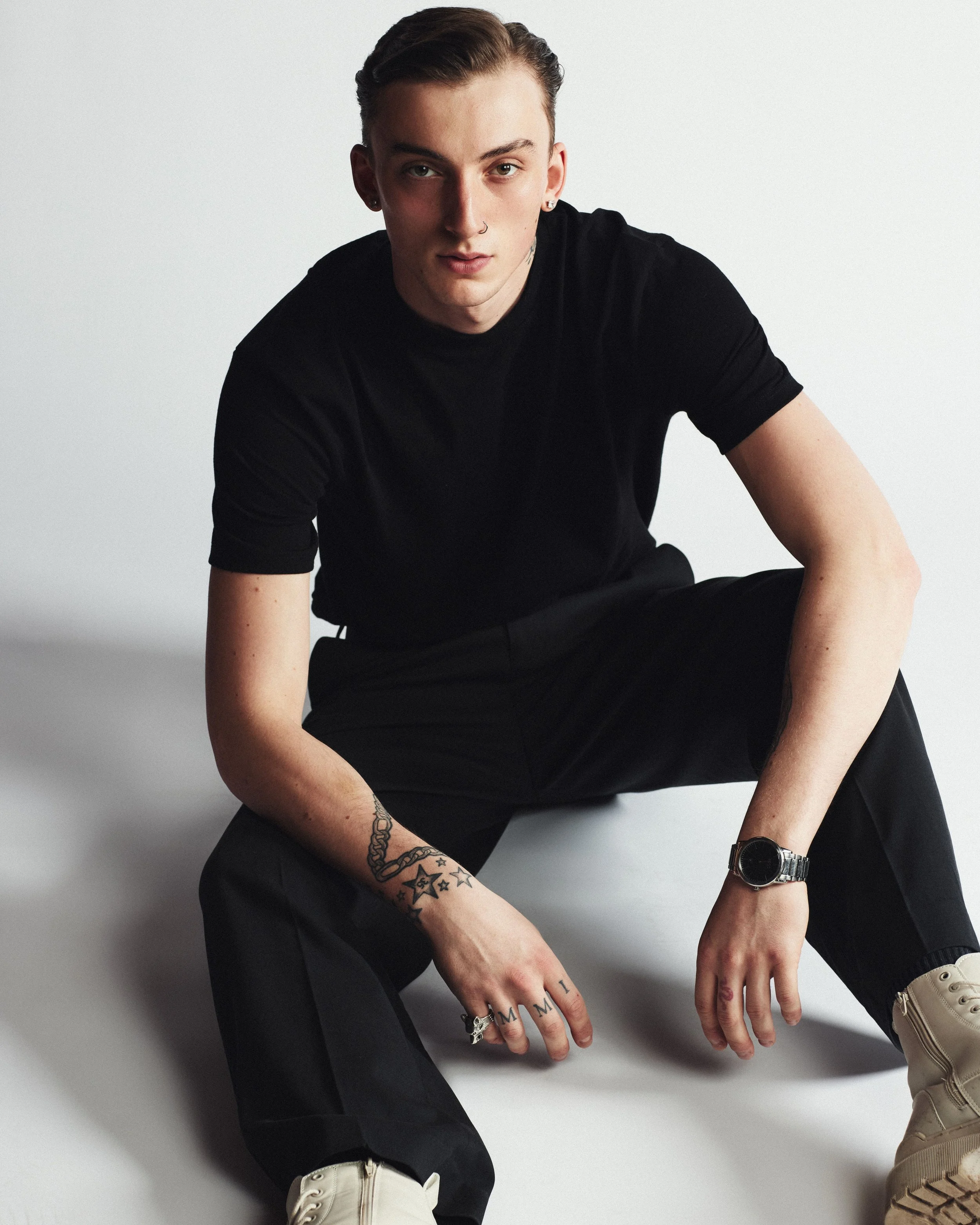 Young man with tattoos on his arm, wearing a black t-shirt, black pants, and beige shoes, sitting on the floor against a plain white background.