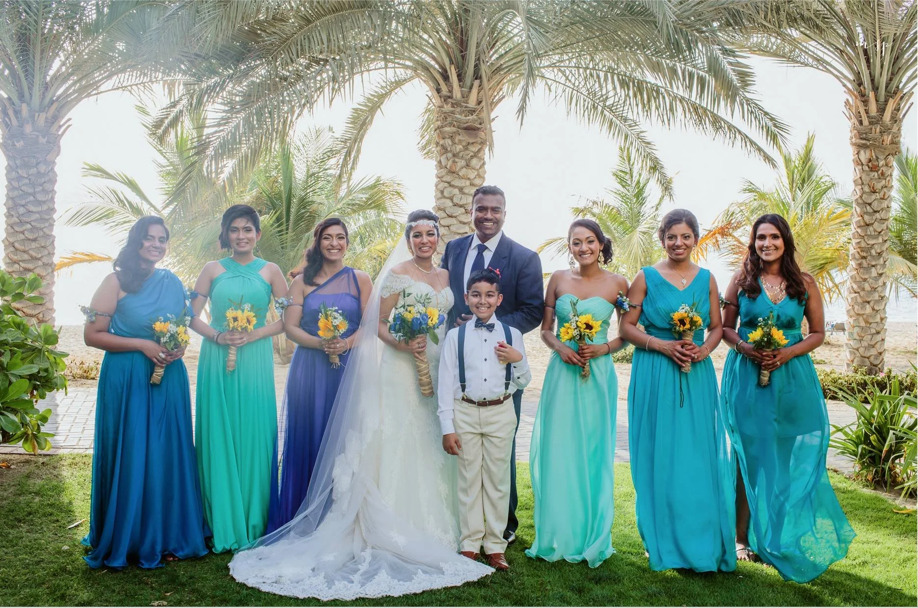 A group of people at a wedding, including a bride in a white wedding gown holding a bouquet, a groom in a dark suit, an excited young boy in a tuxedo, and six women in colorful dresses holding sunflowers, standing outdoors with palm trees in the back