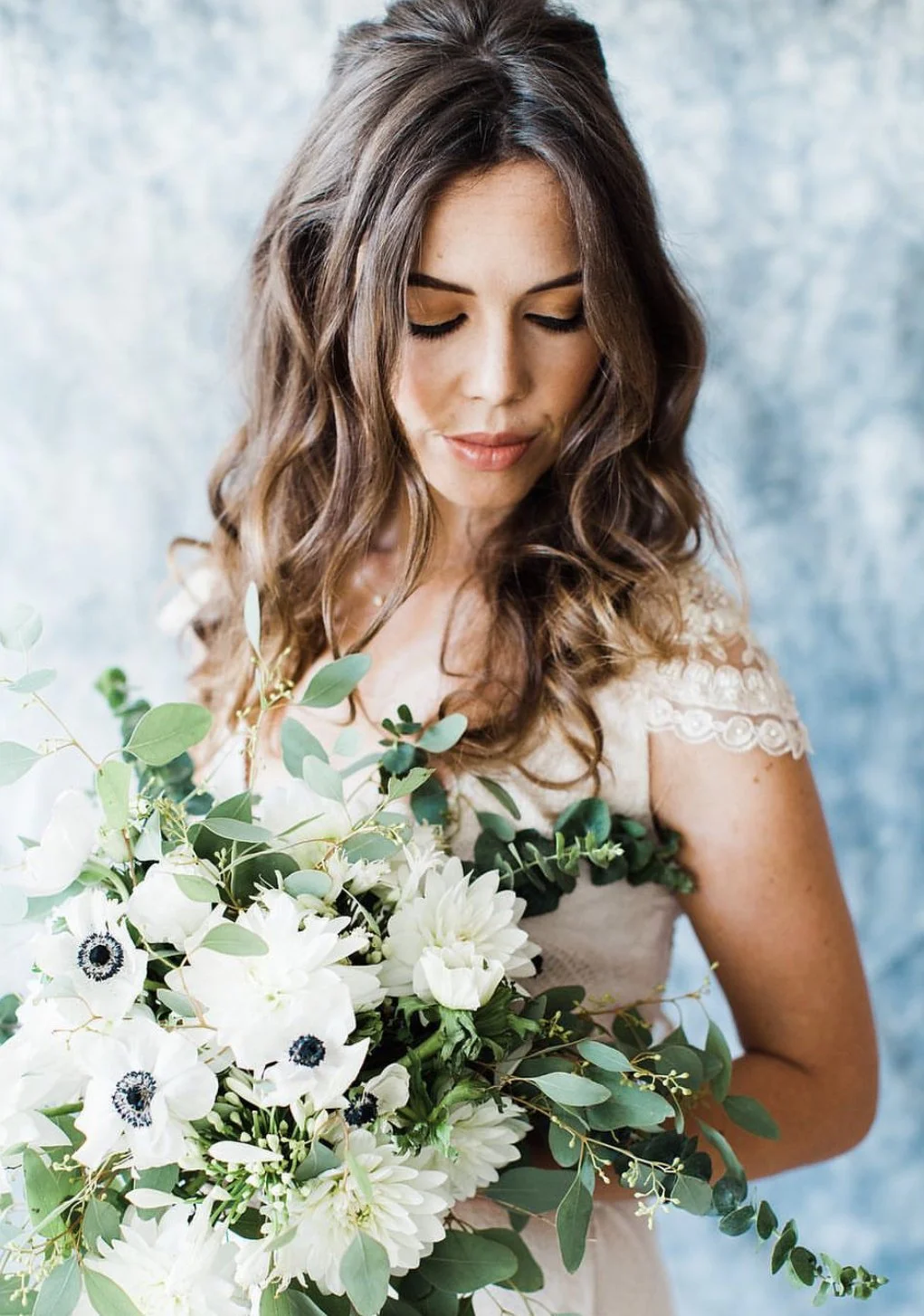 A woman with long, wavy brown hair and closed eyes, holding a large bouquet of white flowers and greenery, in front of a soft blue background.