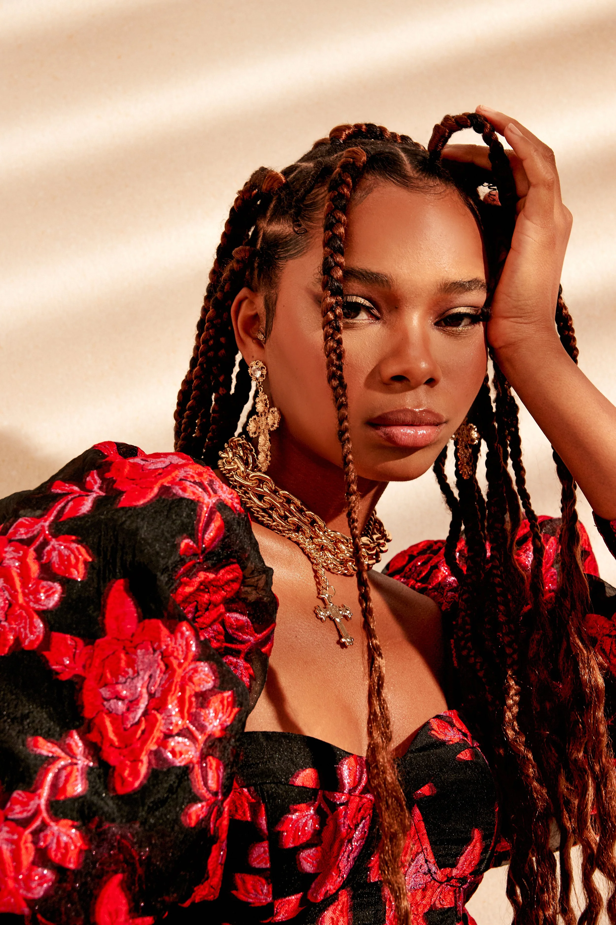A woman with long braided hair wearing a black and red embroidered dress with gold jewelry, striking a pose against a neutral background.