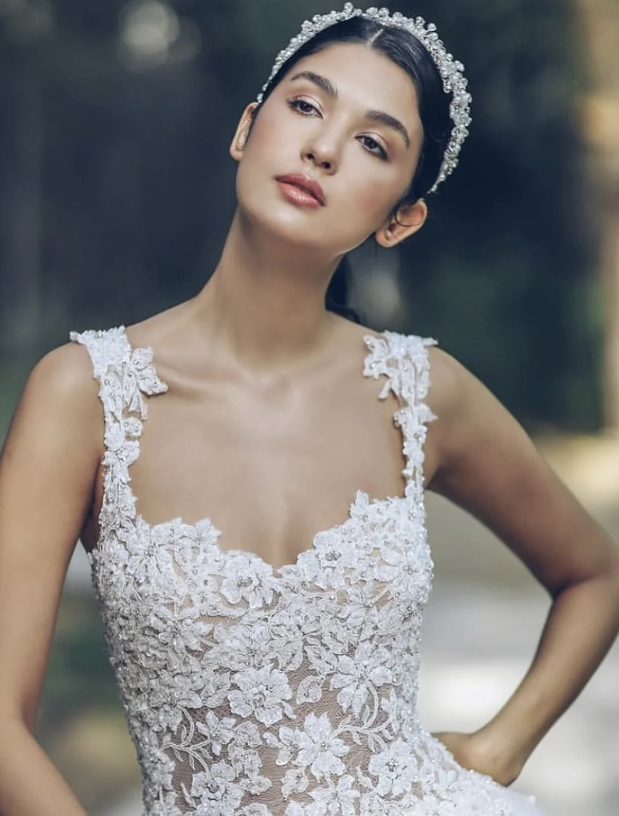 A young woman is dressed in a white lace wedding gown with floral embroidery, wearing a sparkling tiara, outdoors in a natural setting.