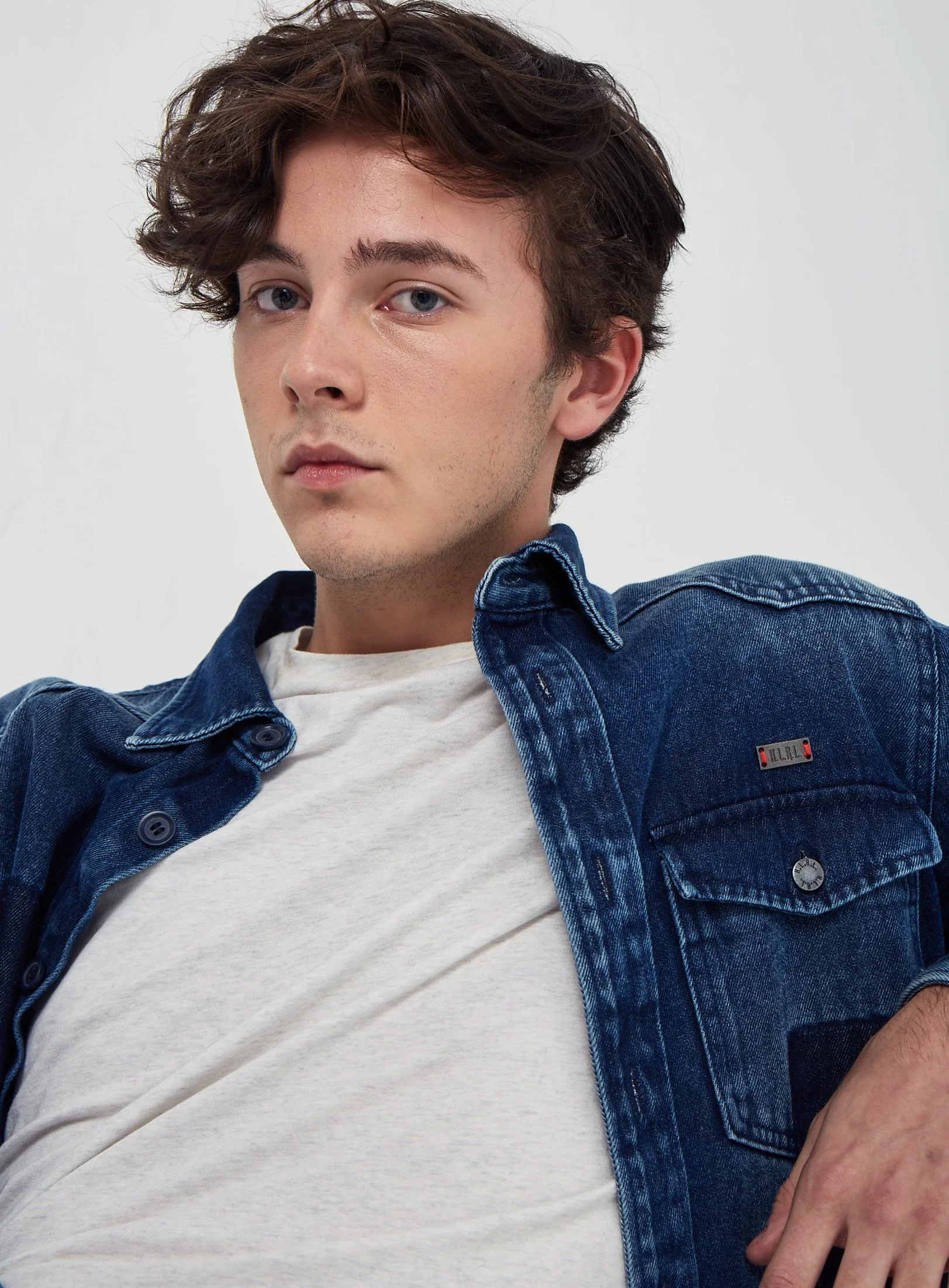 A young man with wavy brown hair and blue eyes posing against a plain white background. He wears a white t-shirt and a dark blue denim jacket.