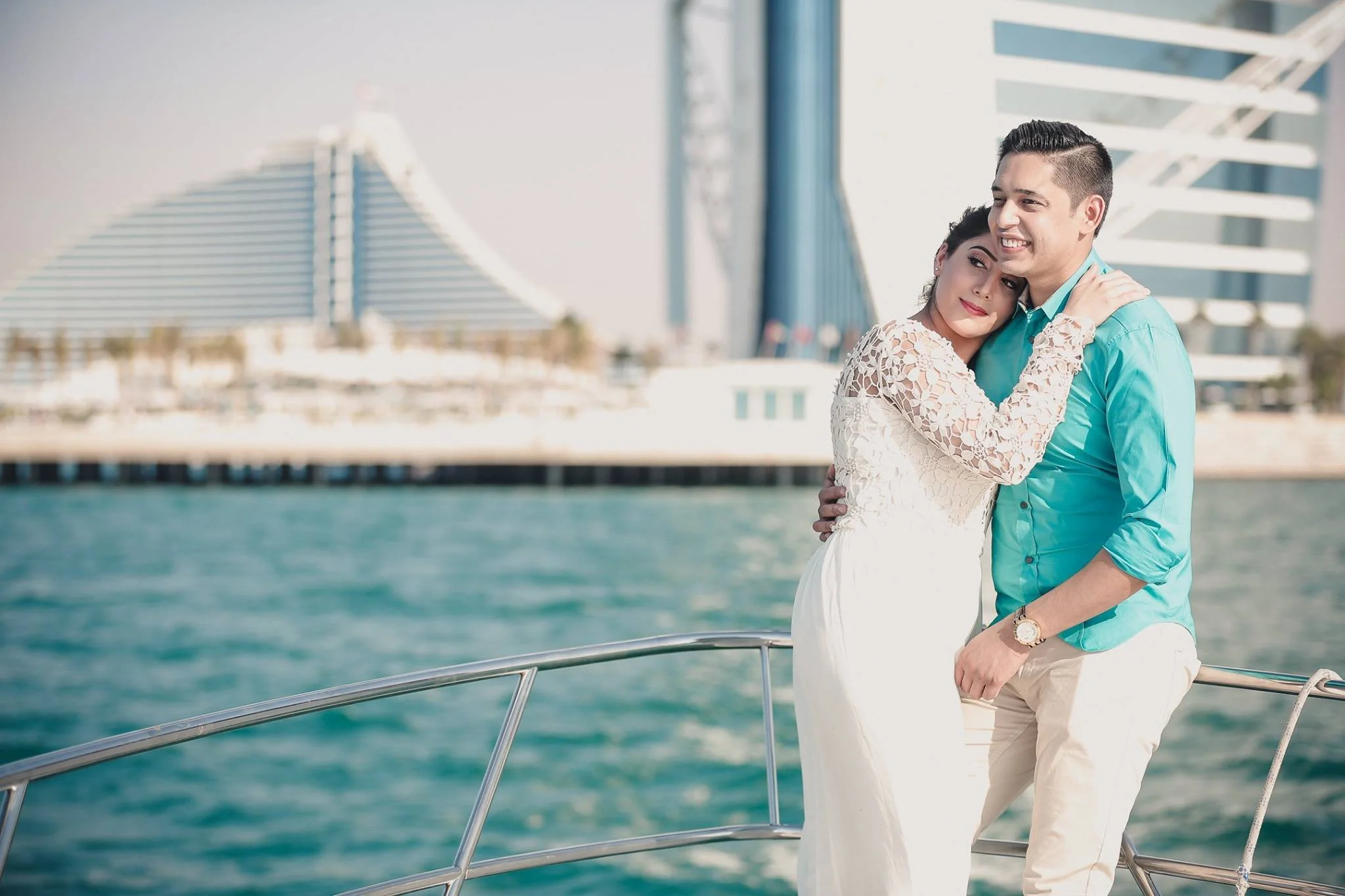 A smiling young couple hugging on a boat with a city skyline and water in the background.