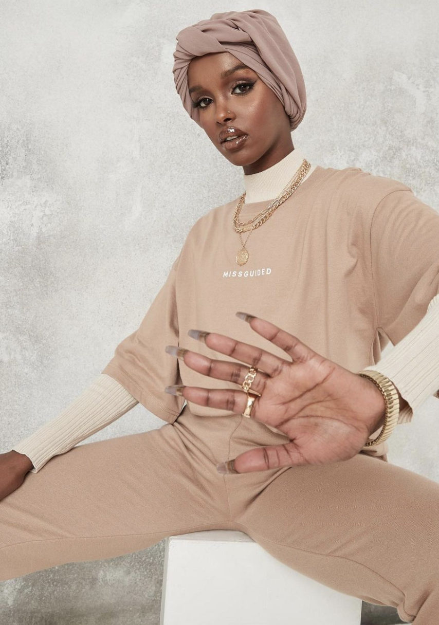 Stylish woman with a beige headwrap, wearing necklaces, rings, and bracelets, sitting with legs apart, gesturing with her hand, against a plain background.