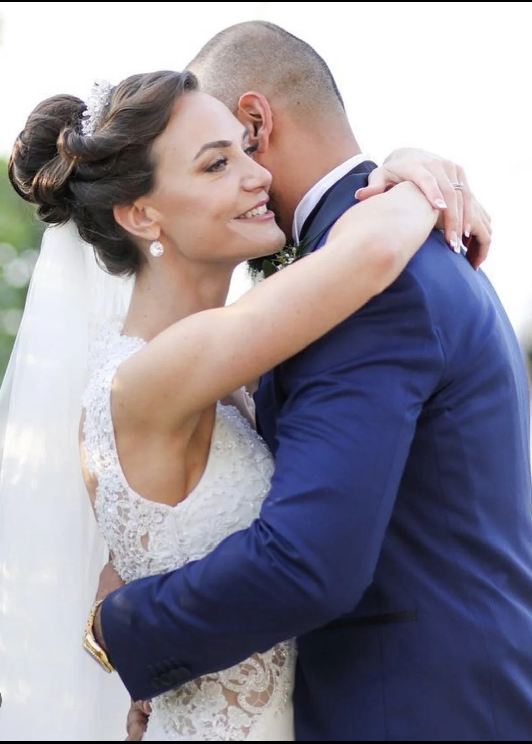 A bride and groom hugging during their wedding, outdoors on a bright day, with the bride smiling and wearing a lace wedding dress and earrings.