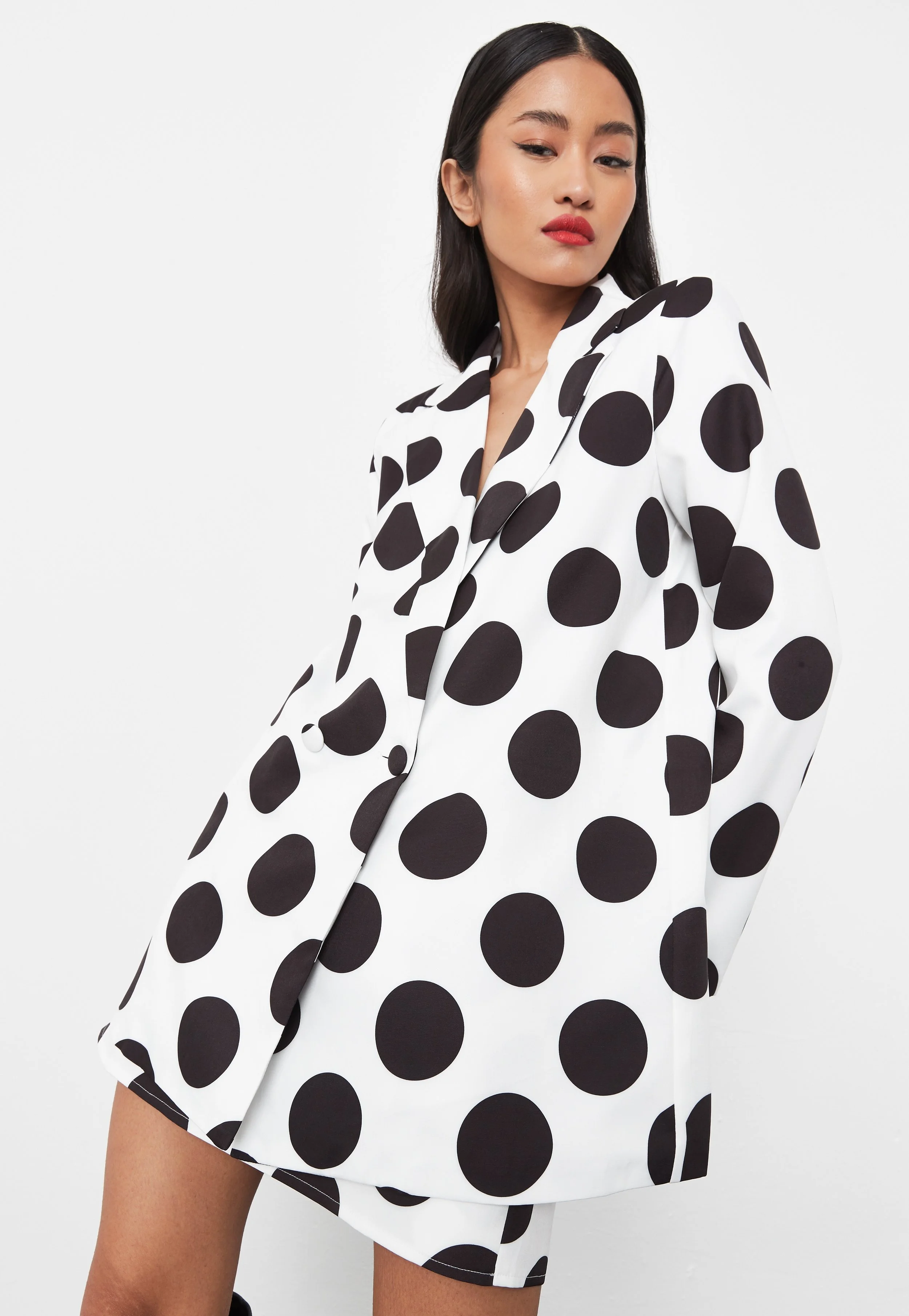 A woman wearing a white blazer and shorts with large black polka dots poses against a plain white background.