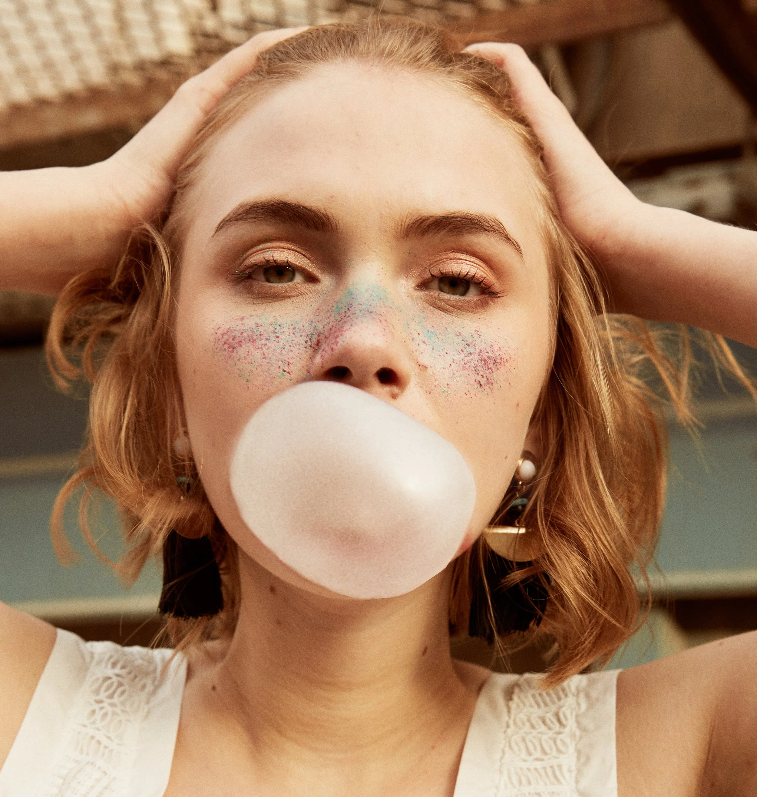 A young woman with light skin and wavy, shoulder-length red hair blowing a bubble with chewing gum, with colorful face paint or glitter on her nose and cheeks. She has her hands on her head, wearing pearl earrings and a white top with lace details.