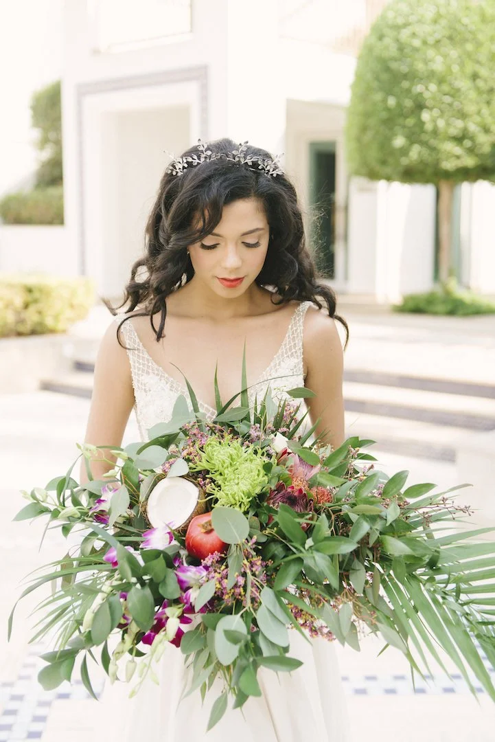 A woman in a wedding dress holding a large bouquet of flowers and greenery outside a building.