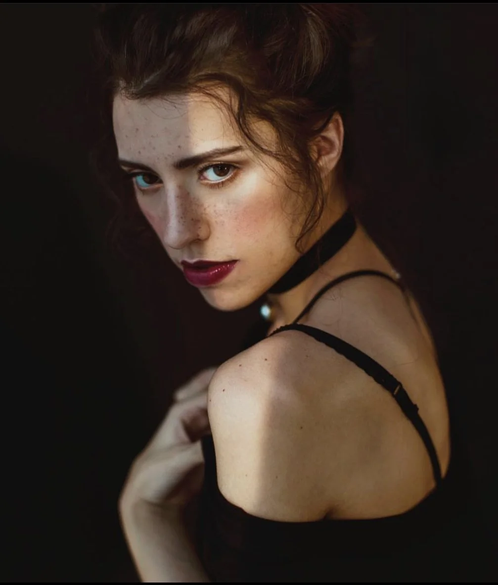A young woman with dark, wavy hair, fair skin with freckles, and striking blue eyes posing against a dark background, wearing a black spaghetti strap top and a choker necklace.