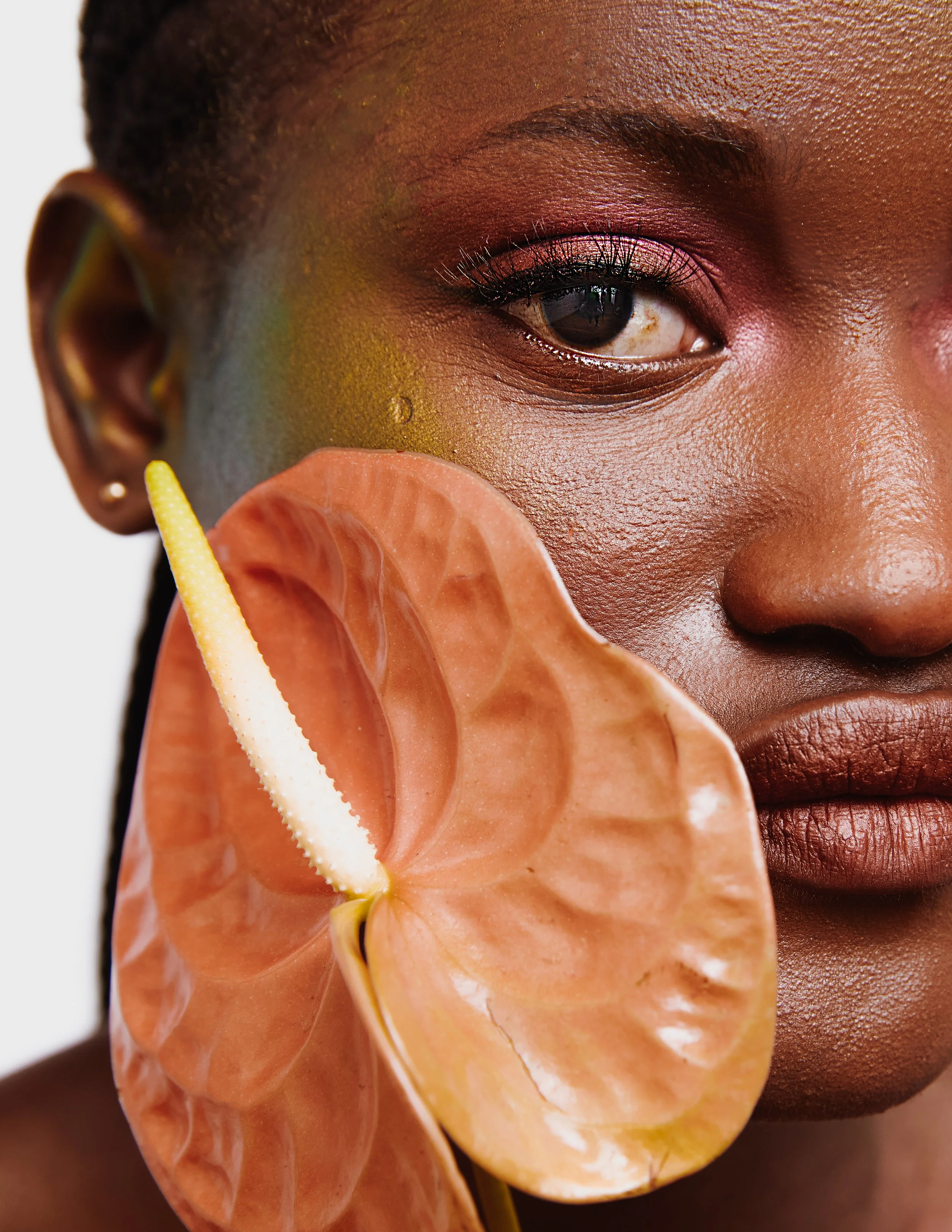 A close-up of a woman's face with colorful makeup and an orange anthurium flower near her cheek.