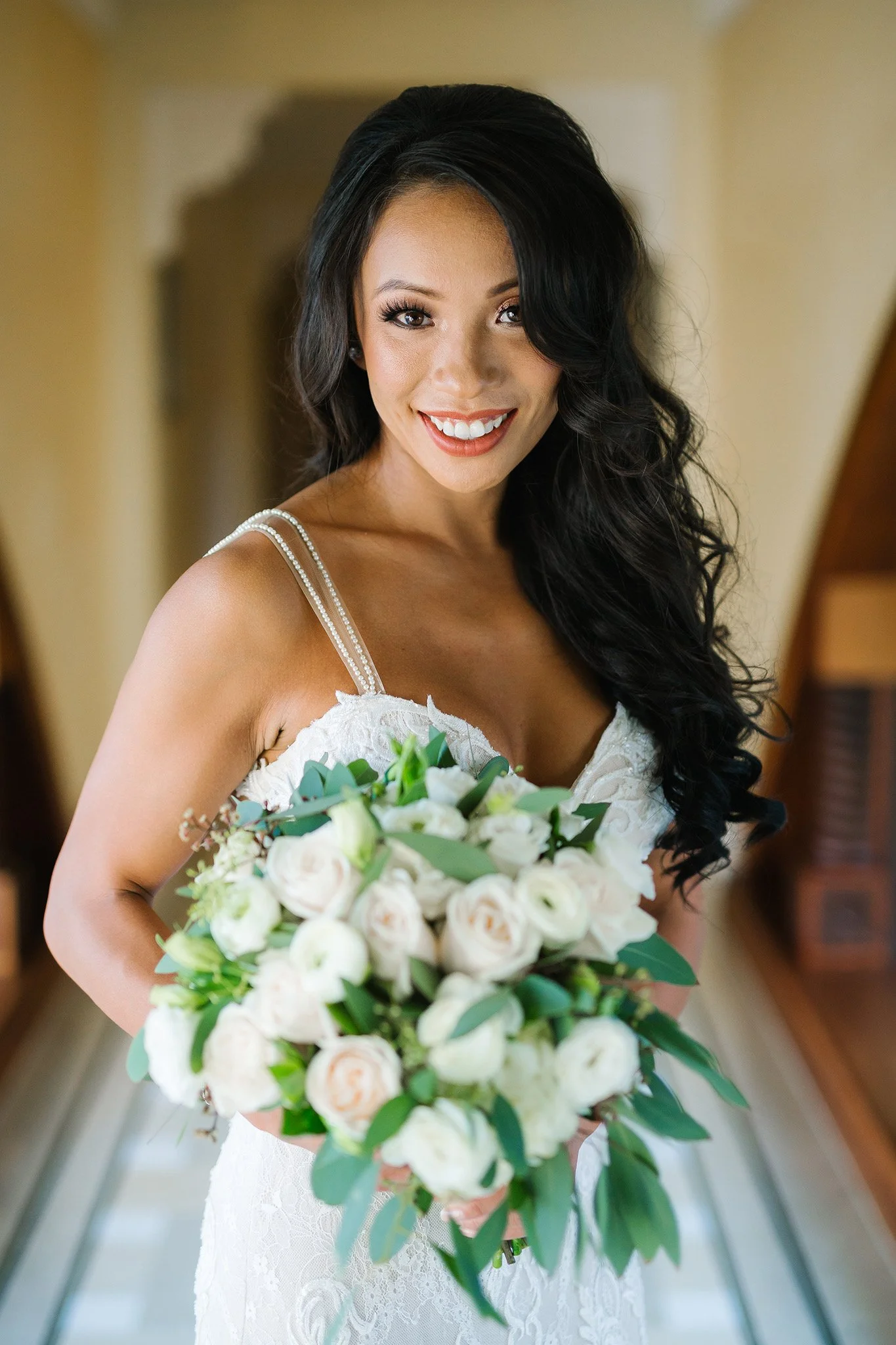 A woman in a white wedding dress holding a bouquet of white and green flowers, smiling at the camera.