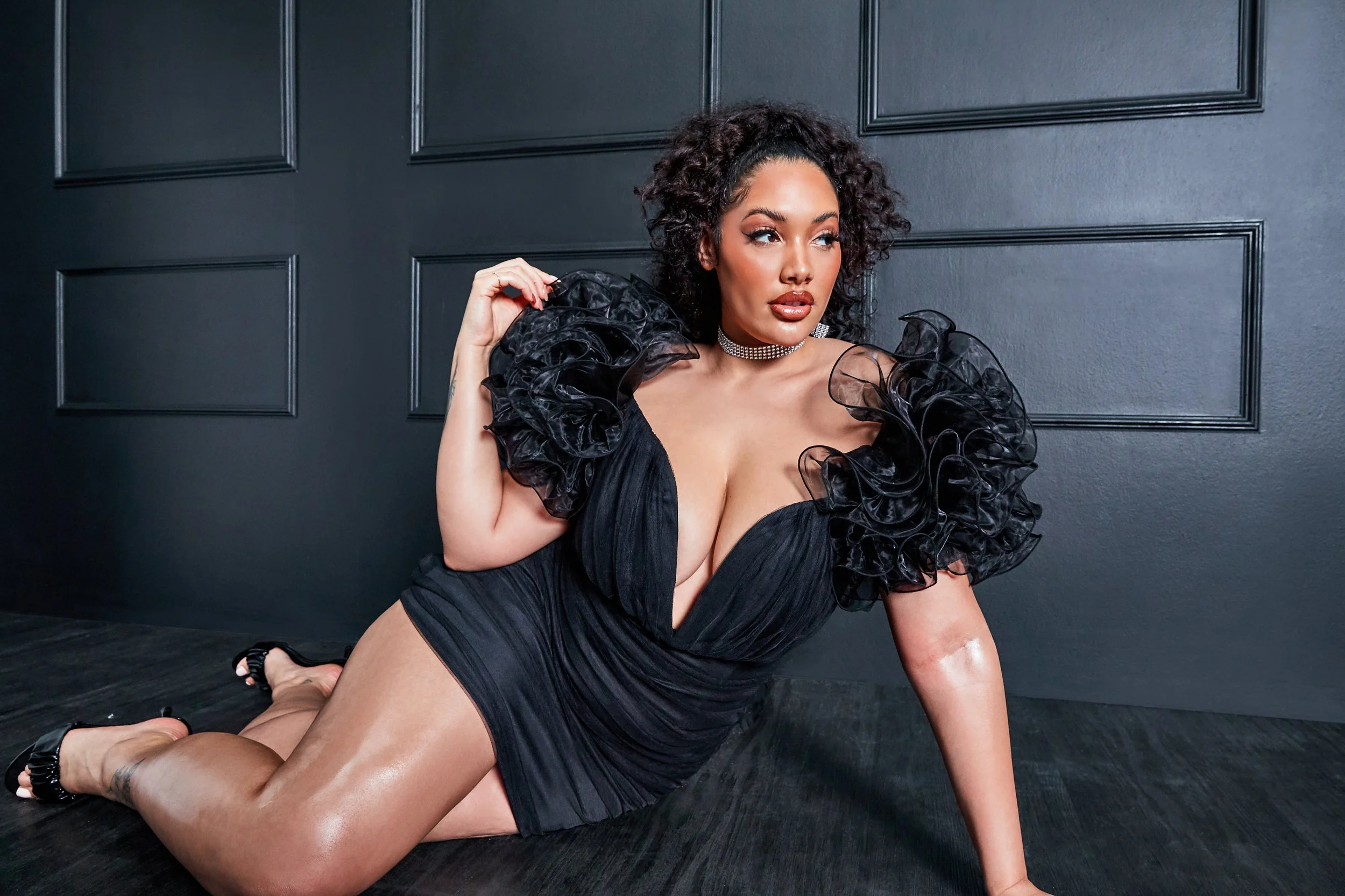 A woman in a black dress with ruffled sleeves is posing on a dark floor against a dark wall with decorative panels.