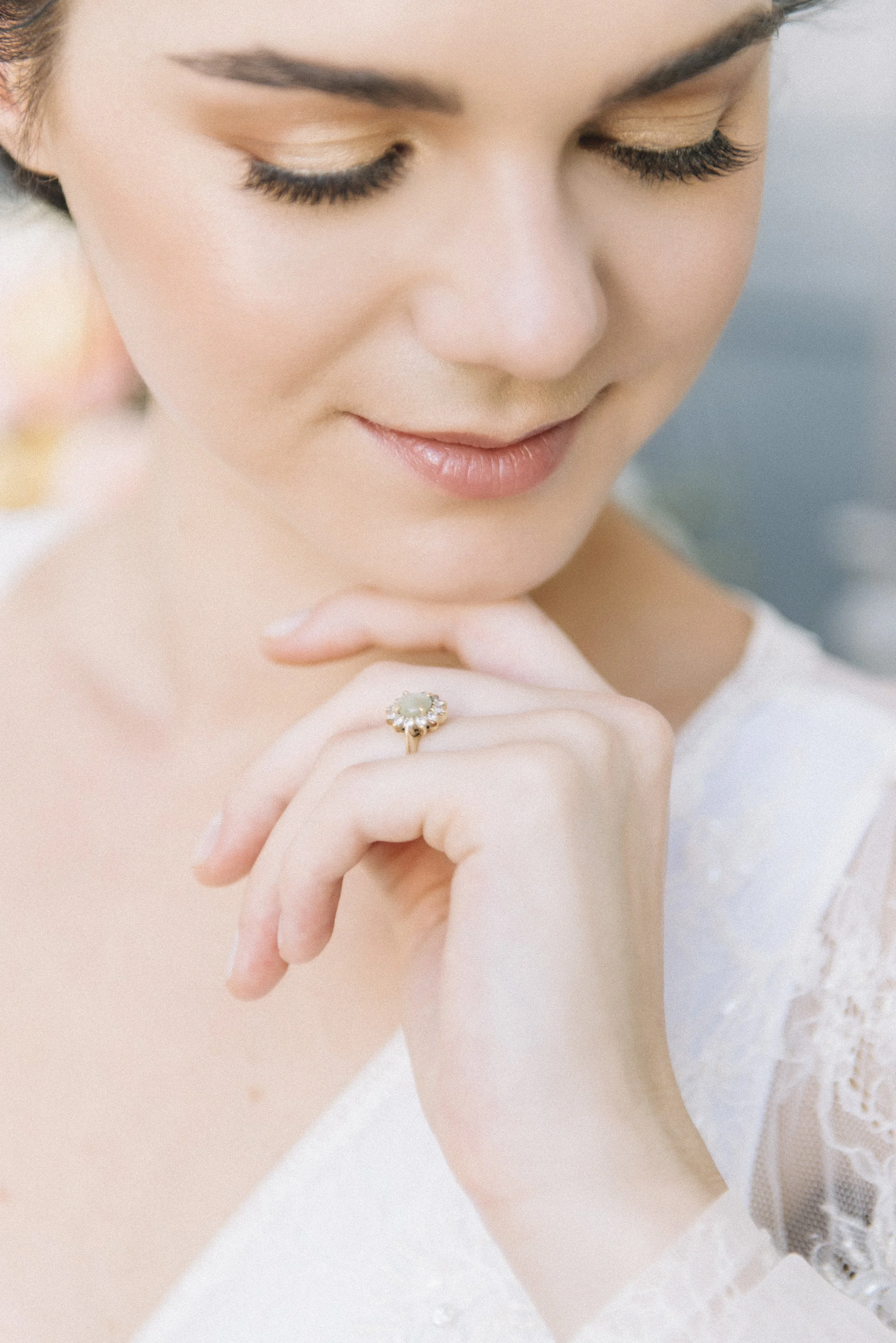 Close-up of a woman with closed eyes, wearing light makeup, a pearl engagement ring, and a white lace dress.