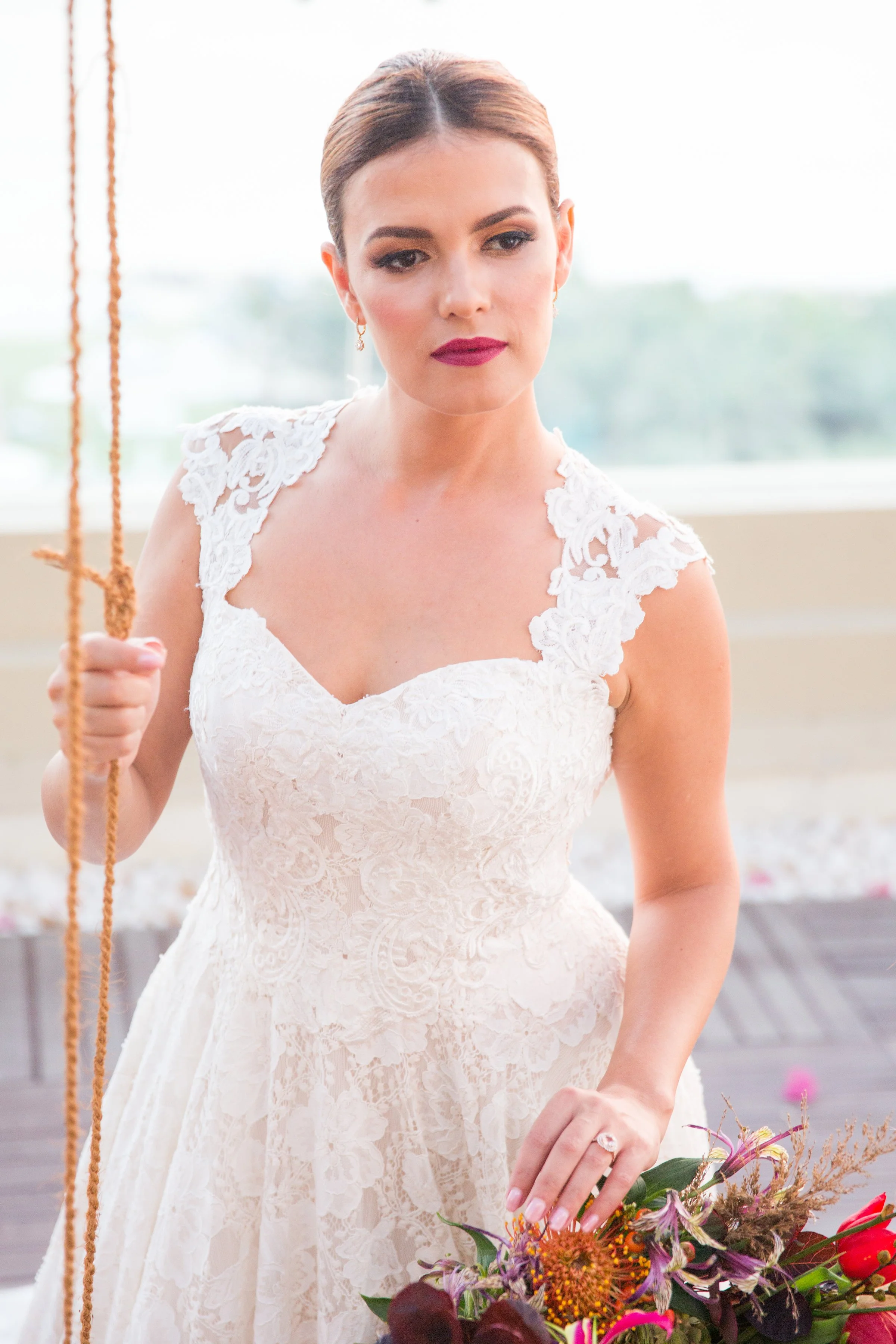 A woman wearing a white lace dress, holding a bouquet of flowers, standing indoors with a blurred window background.