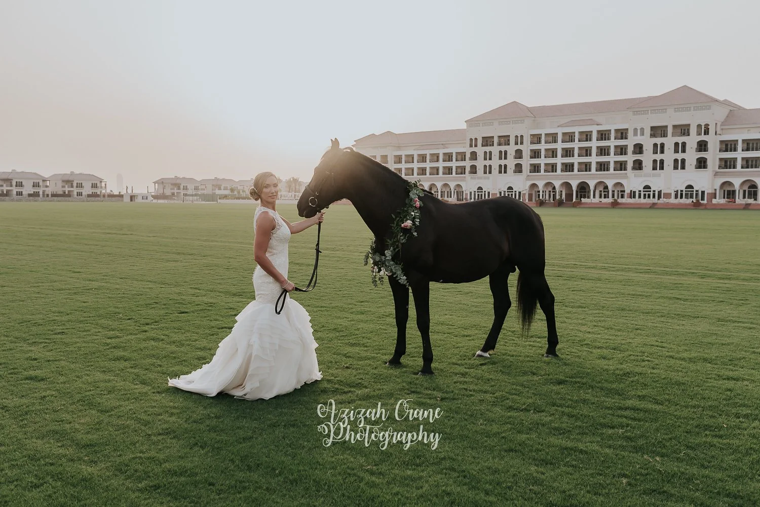 Bride in a white wedding gown holding a black horse with a floral wreath at a green field with a large white building in the background.