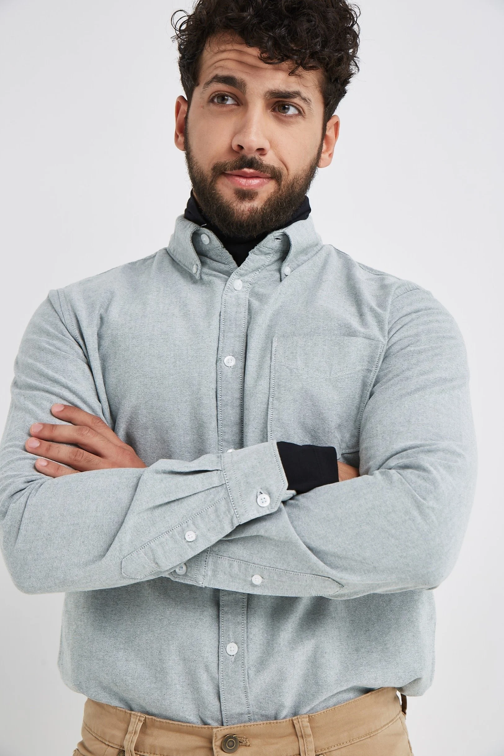 A man with dark curly hair and a beard, wearing a light gray button-up shirt over a black turtleneck, crossing his arms and looking to the side against a plain white background.