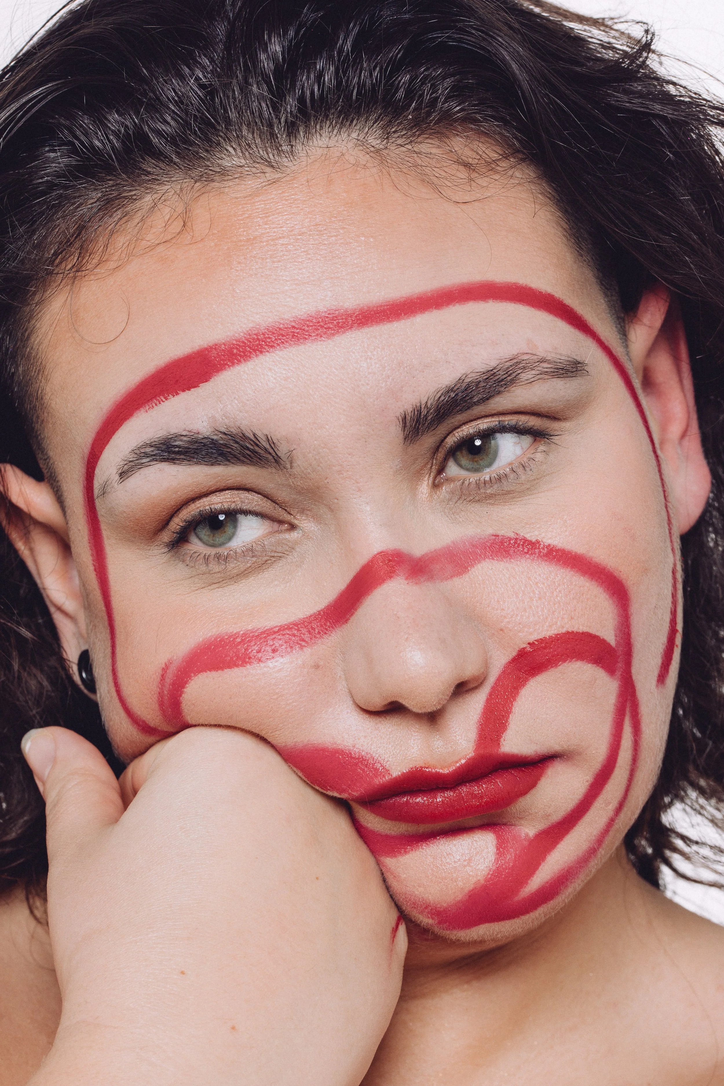 Close-up of a person's face with red lipstick and red lines painted across their face, resting their chin on their hand.