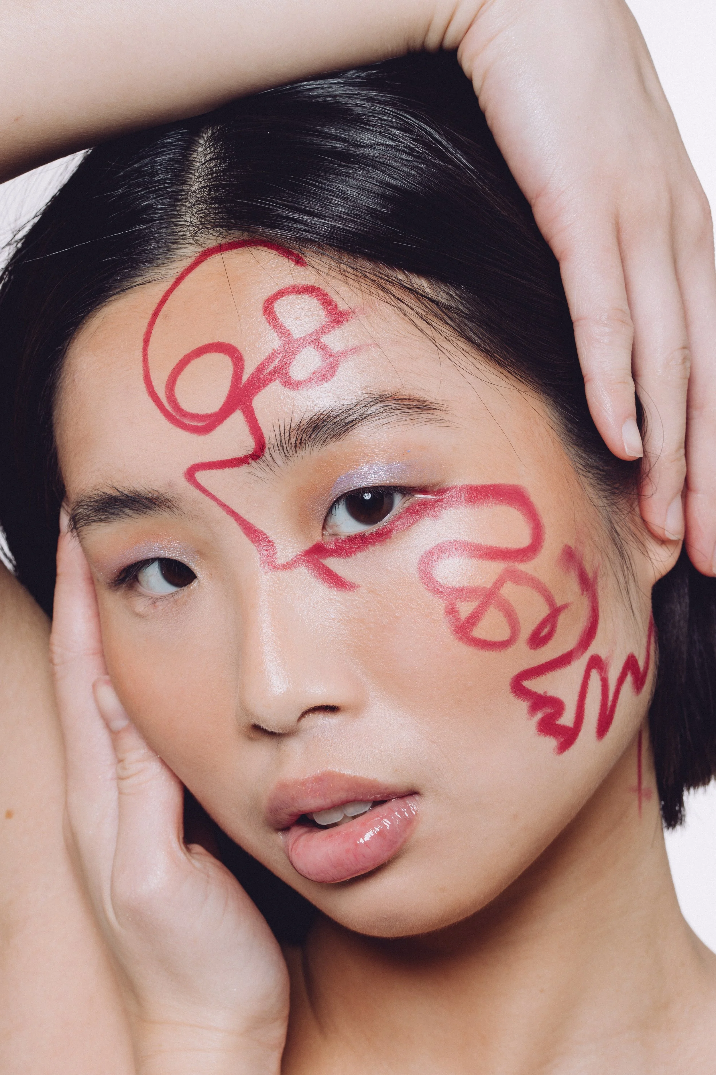 Close-up portrait of a young woman with short black hair, light makeup, and red lipstick. She has red graffiti-style writing on her face and head, and her hand is resting on her head, framing her face.