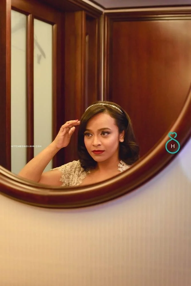 A woman with shoulder-length dark hair, wearing a dress with lace details, is looking into a mirror while adjusting her hair, with a wooden door and paneling in the background.
