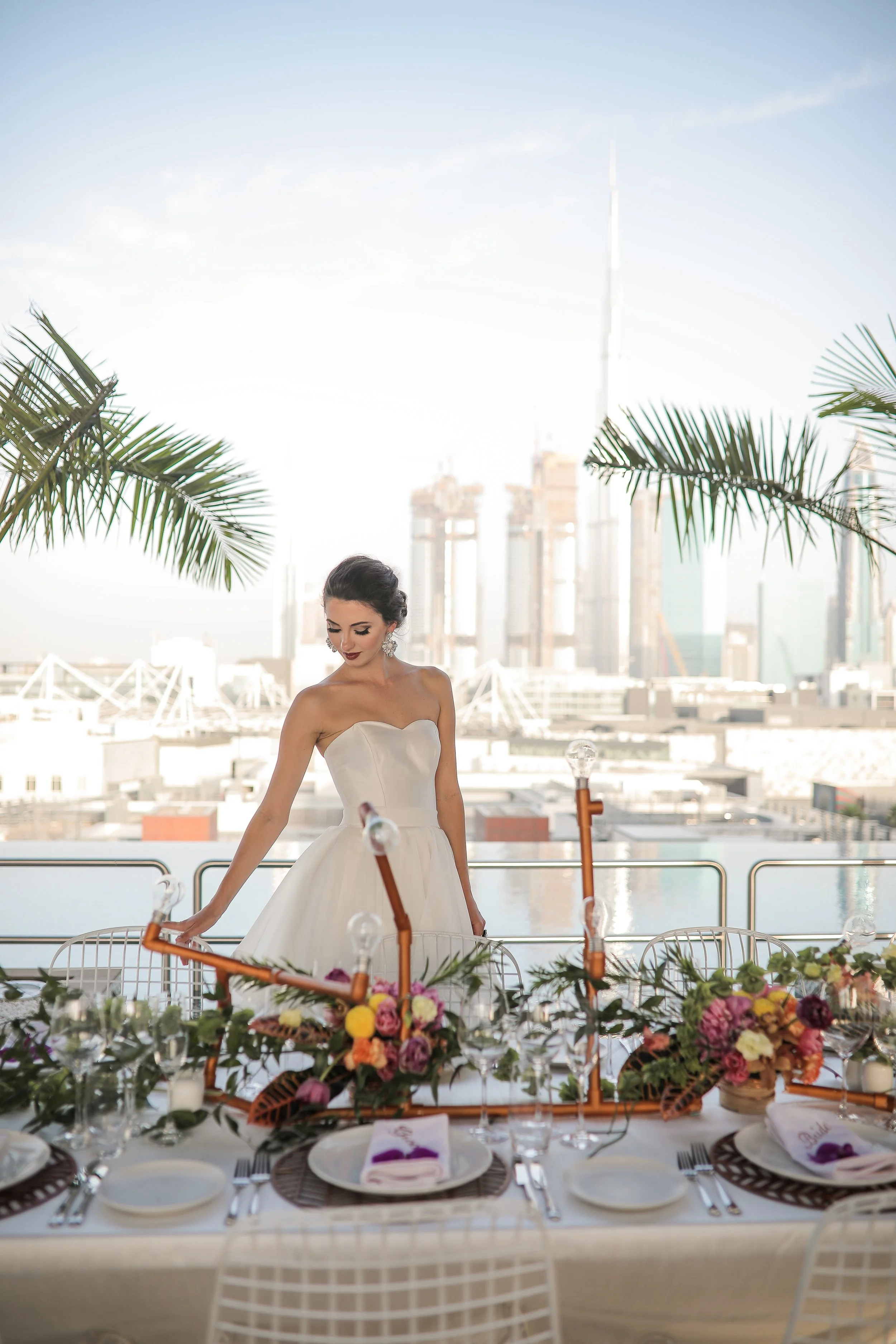 A woman in a strapless white wedding dress stands at a decorated dining table on a balcony with a city skyline in the background.