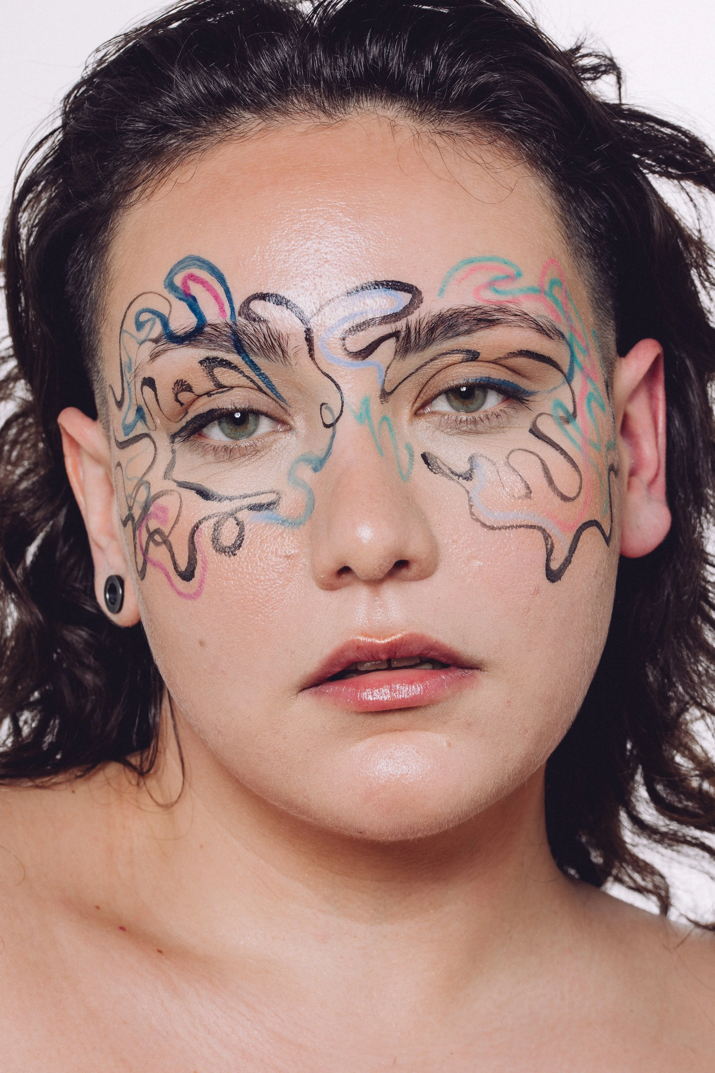 A close-up of a person with light skin and curly dark hair, makeup, and colorful face paint designs over their eyes and cheeks.