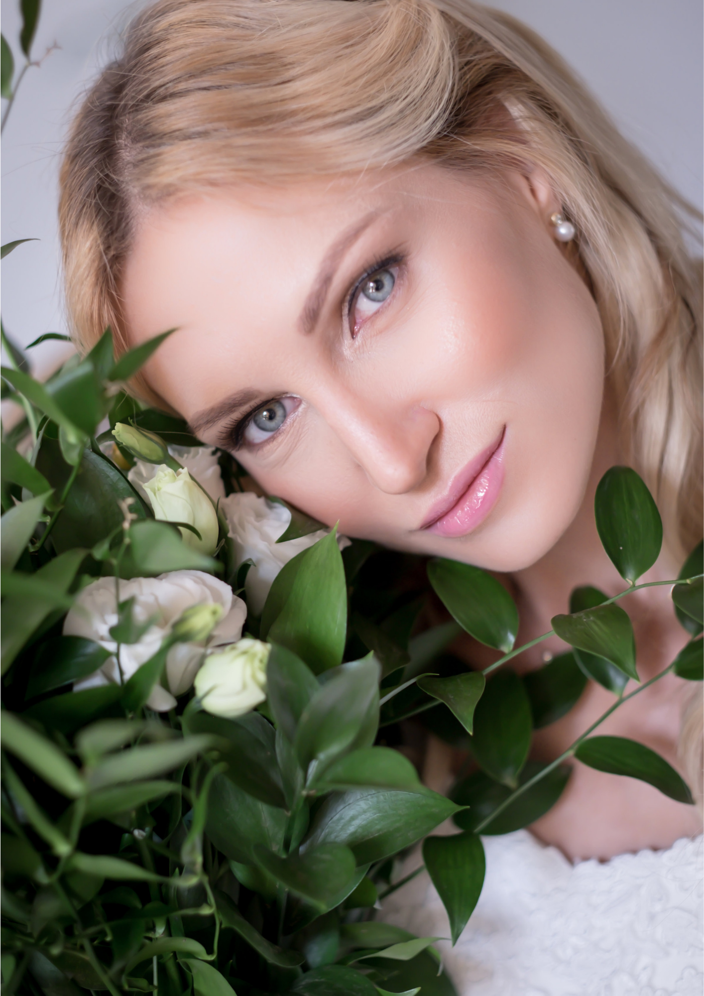 A woman with blonde hair and blue eyes resting her face against white flowers and green foliage.
