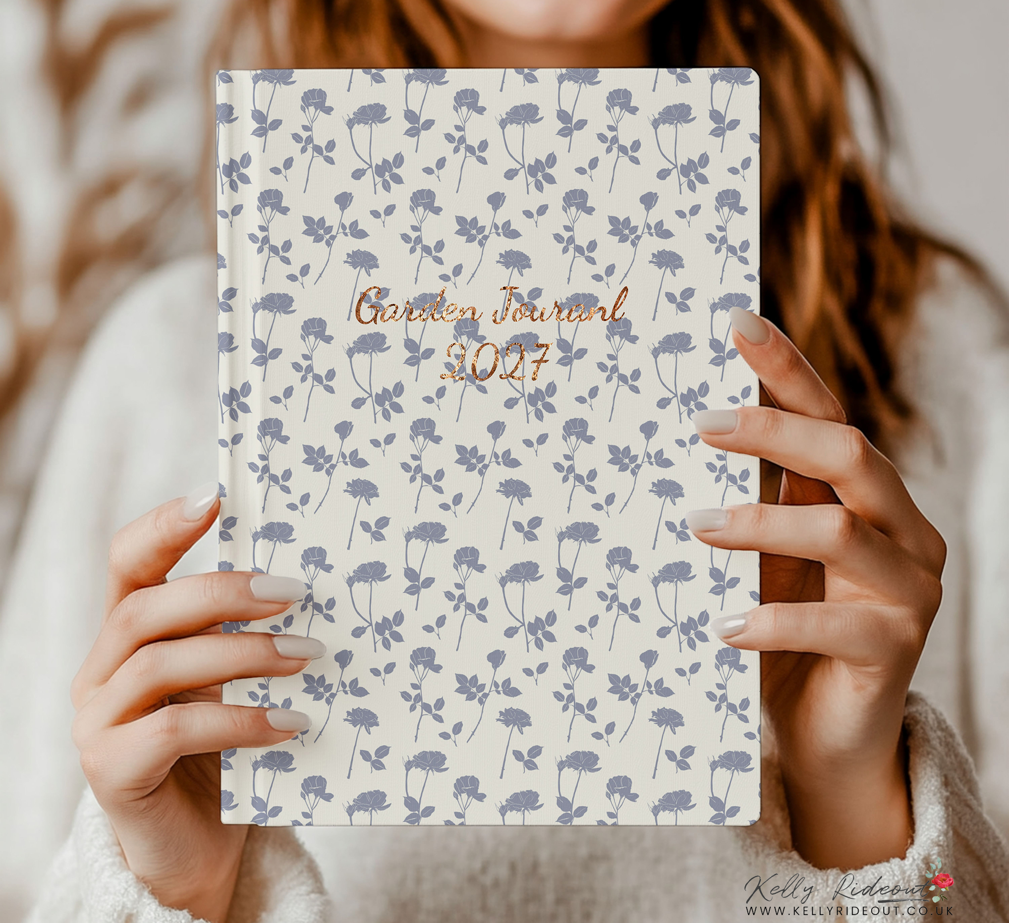 A person holding a garden journal with a floral pattern cover, the text 'Garden Journal 2027' written in script font, with a person wearing a white sweater. 