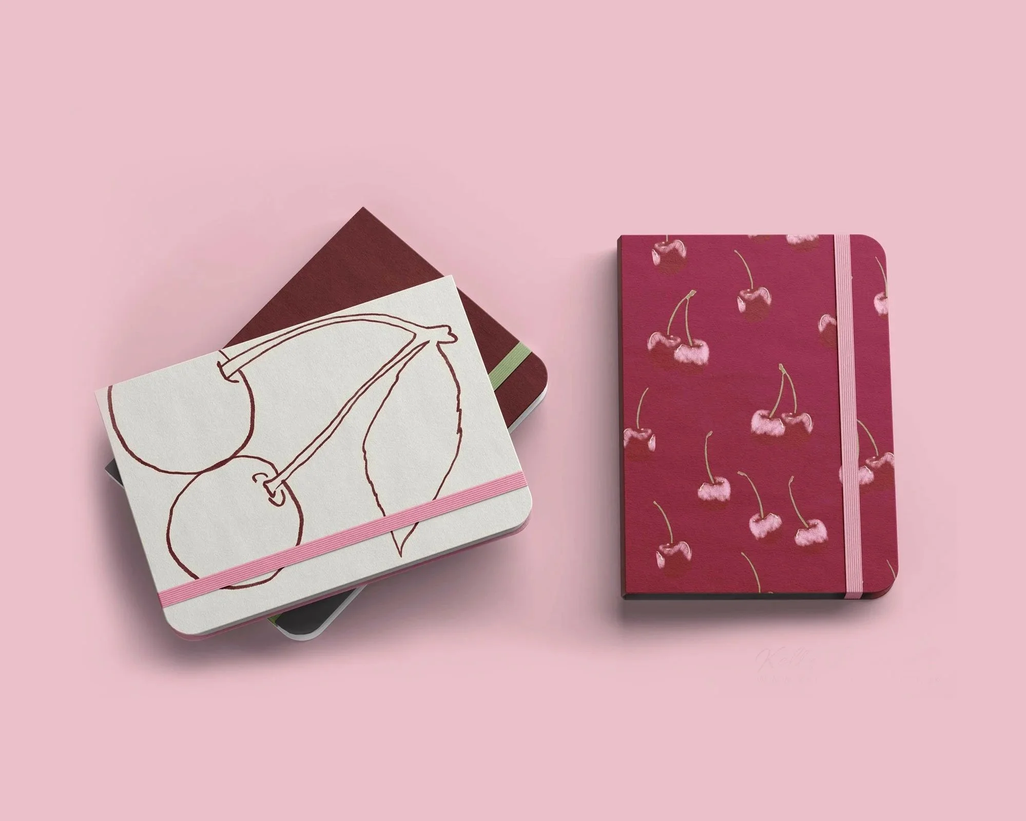 A flat lay of two closed notebooks, one with a cherry pattern and pink elastic band, and the other with a plain maroon cover, on a pink background. Designed for art licensing by Kelly Rideout