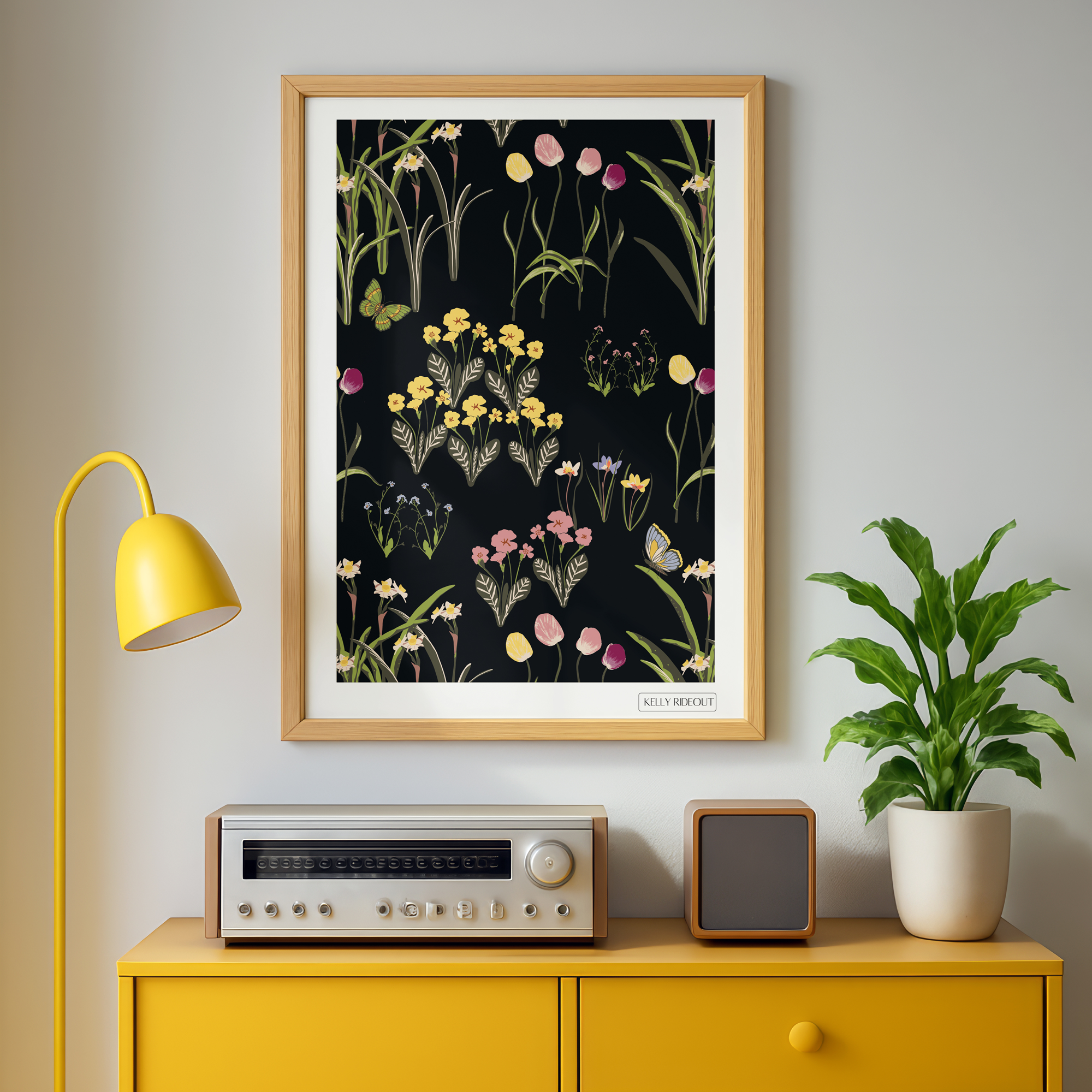 Spring floral wall-art print on dark background by Kelly Rideout A framed botanical artwork with flowers and butterflies hangs on a neutral wall above a yellow console table with an electronic device, a potted plant, and a yellow desk lamp nearby.