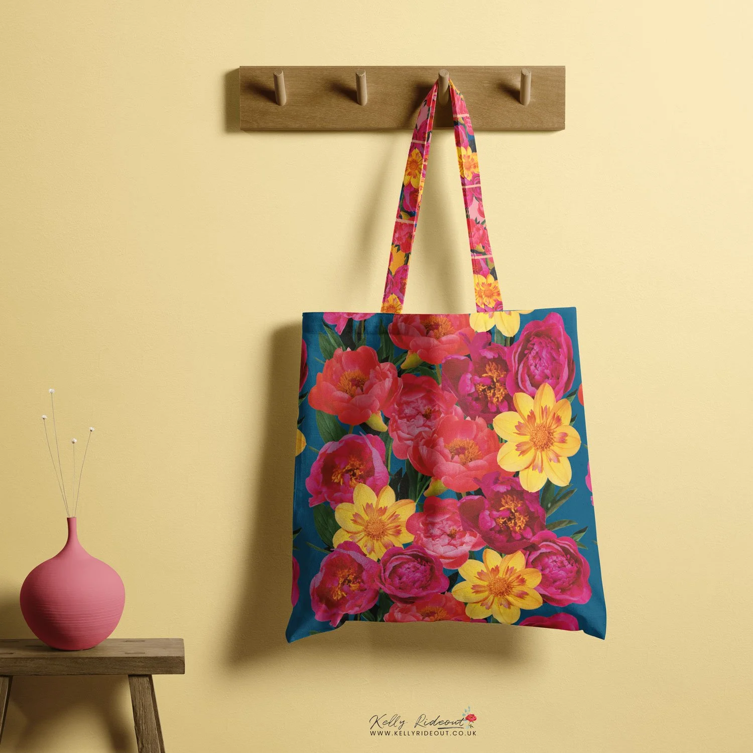 Colorful floral tote bag hanging on a wooden hook on a yellow wall, with a small pink vase on a wooden table to the left. Example of Kelly Rideout's patterns for licensing  on product