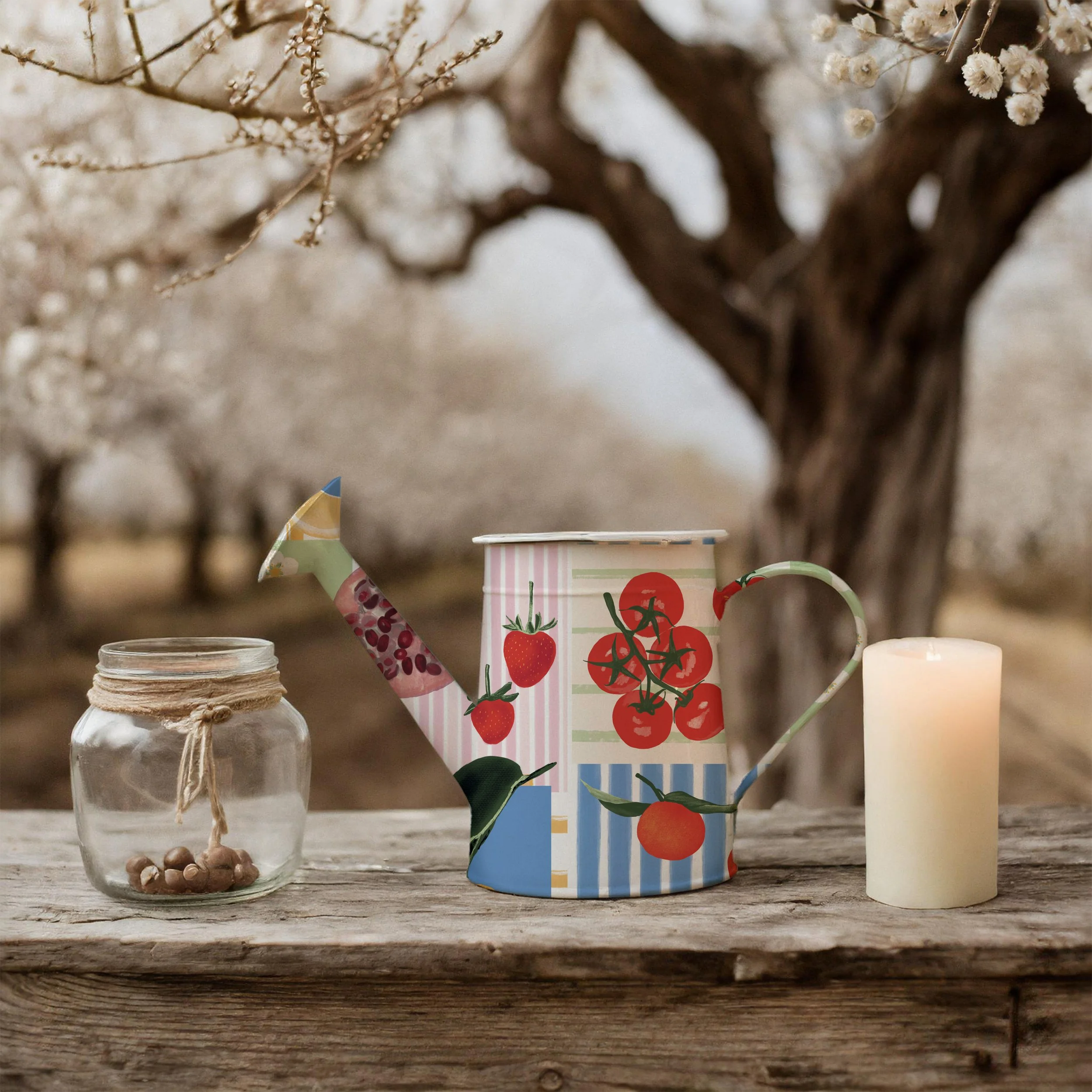 A decorative watering can with fruit illustrations, a glass jar with a string tie and small nuts inside, and a white candle on a rustic wooden surface outdoors with blooming trees in the background.