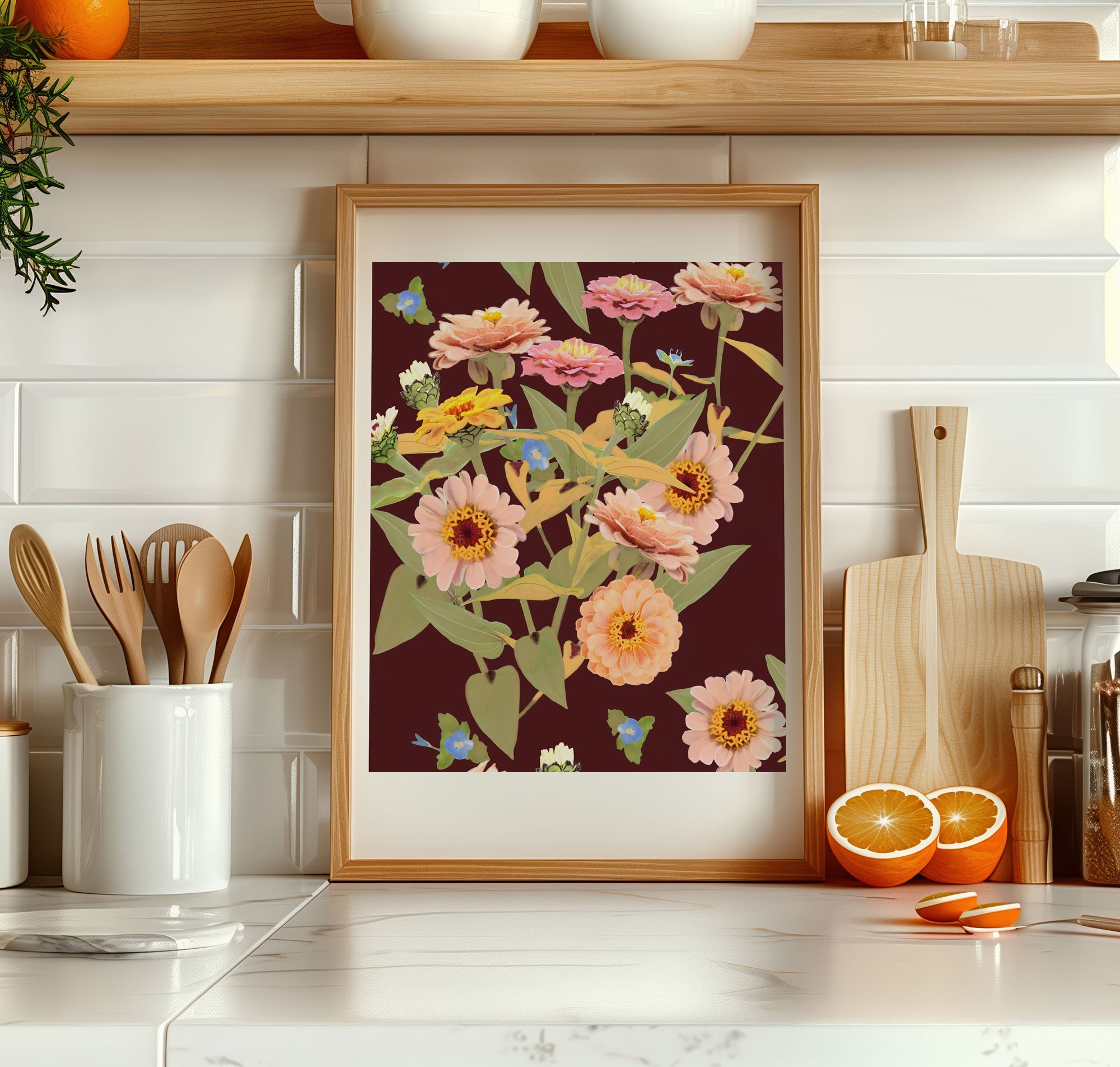 A framed floral illustration with pink and yellow flowers and green leaves, placed on a kitchen counter with wooden spoons, a cutting board, halved oranges, and other kitchen items, with a white tiled wall background.