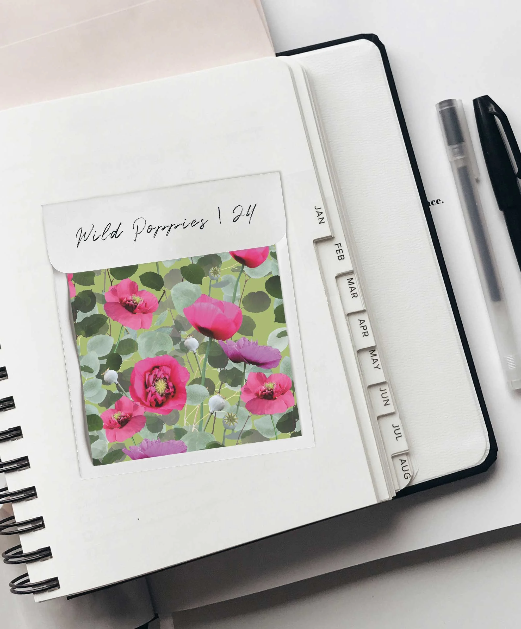 Open planner with a note labeled "Wild Poppies 24" and a picture of pink poppies beneath it. The planner has tabs for each month, with visible months including January, February, March, April, May, June, July, and August. Two pens are placed to the r