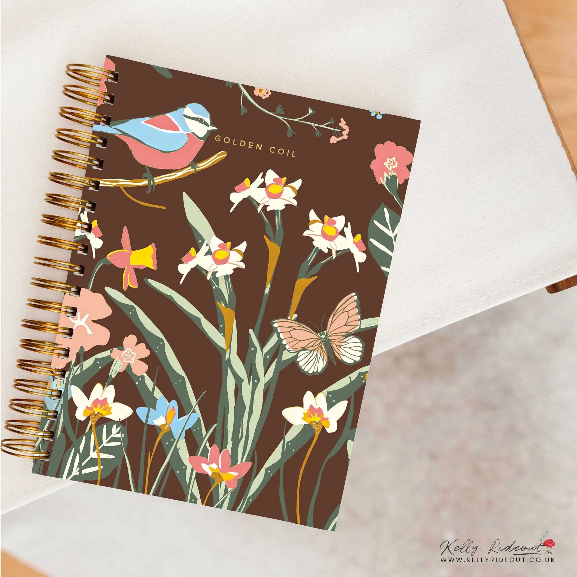 A decorated spiral notebook with a floral and bird design on the cover, featuring a blue and pink bird, colorful flowers, and a butterfly, placed on a white surface. Design for Golden Coil competition