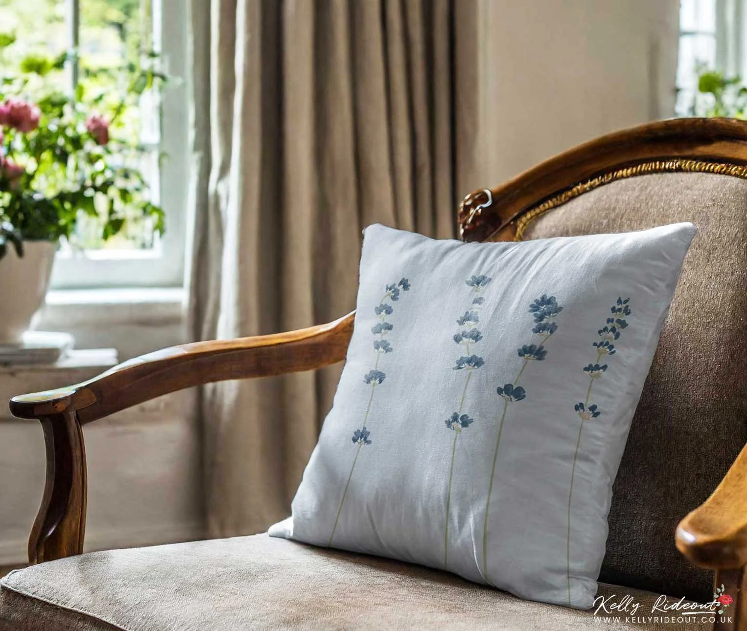 A light blue pillow with blue lavender embroidery resting on a vintage wooden armchair with beige upholstery, in a well-lit room with curtains and a window showing greenery outside. Lavender illustration for licensing for home decor