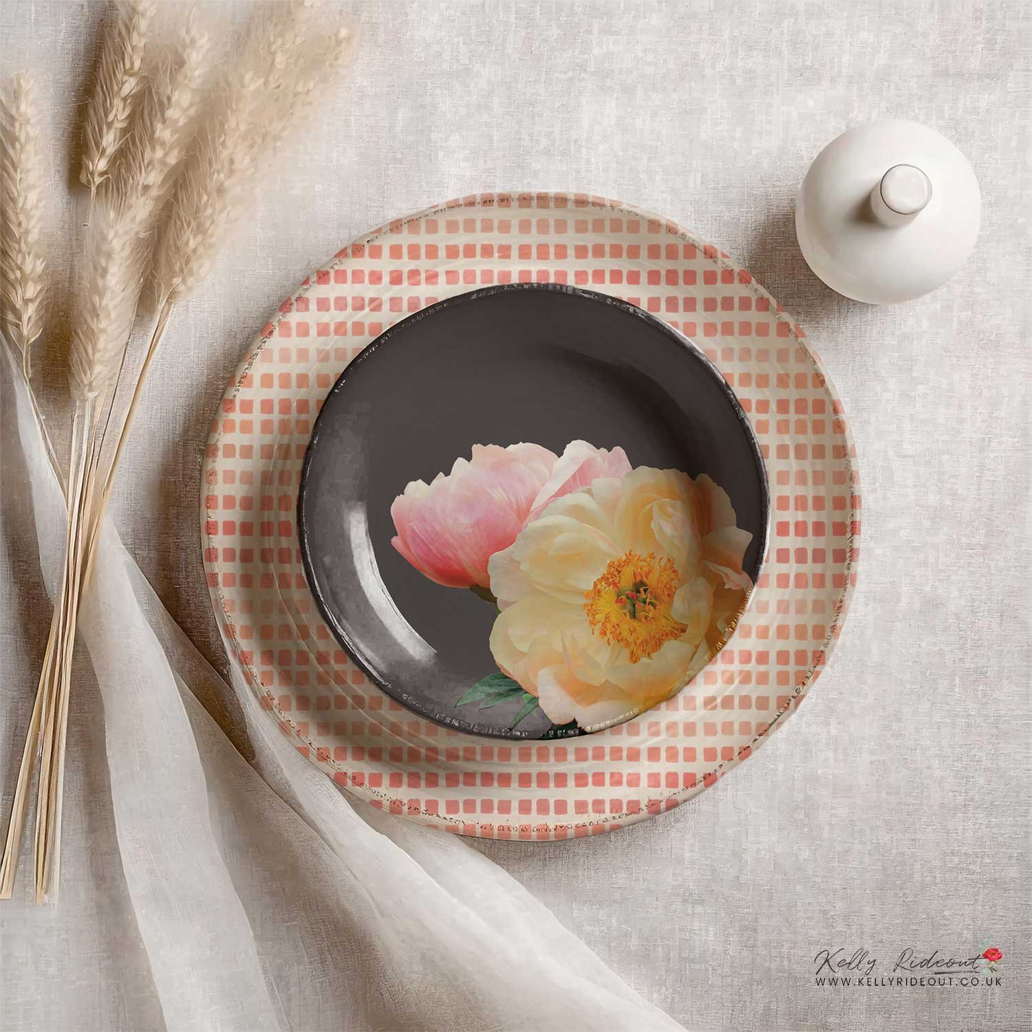 A layered place setting on a cream tablecloth featuring a pink and white checkered large plate, topped with a smaller Silhouette colour bowl with floral design, pattern designs.