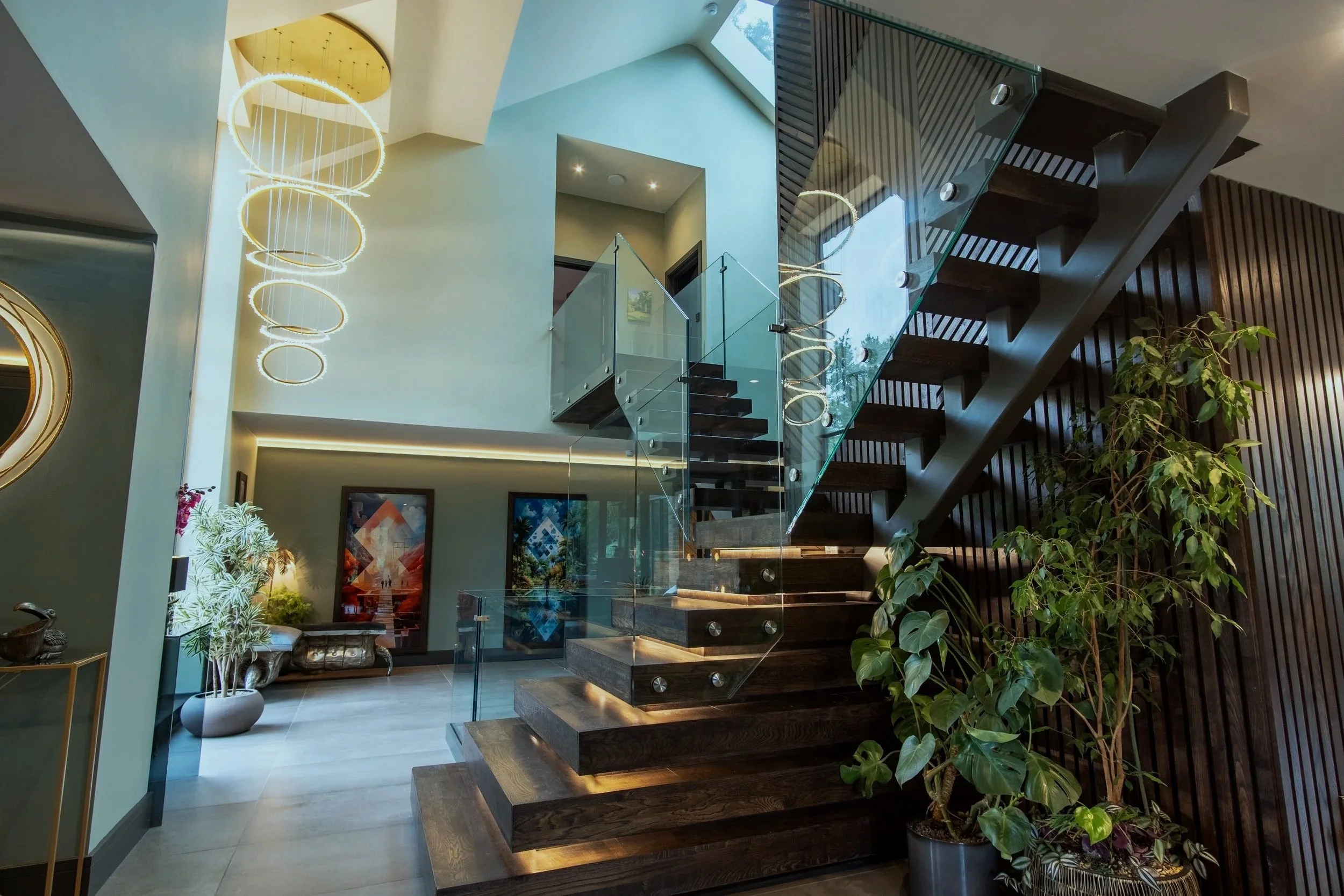 Modern interior with a glass staircase, wooden steps, and contemporary lighting fixtures. The space includes decorative wall art, plants, and a sitting area.