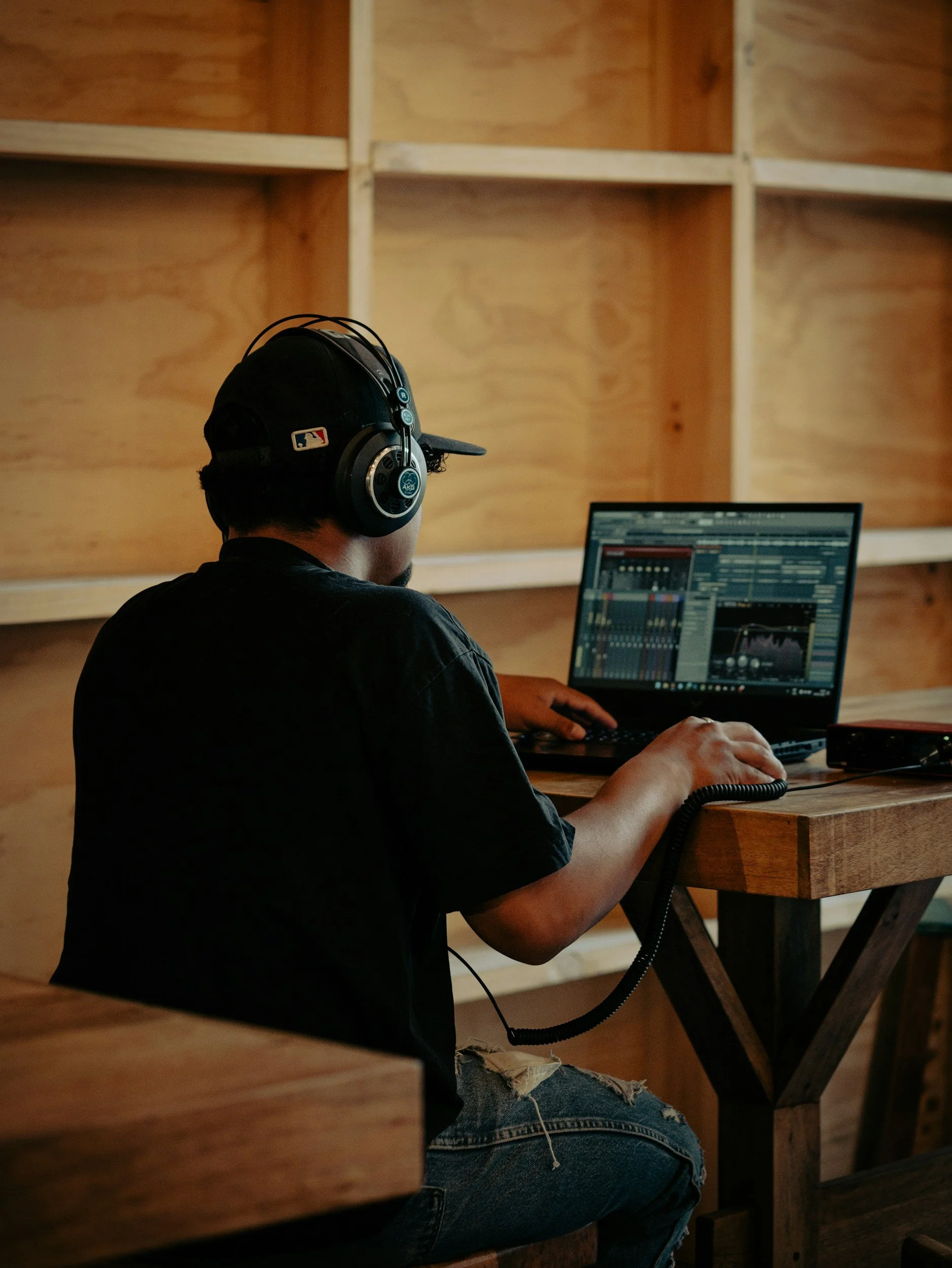 A person wearing a black baseball cap, headphones, and a black T-shirt working on a music production software on a laptop in a wooden studio or room.