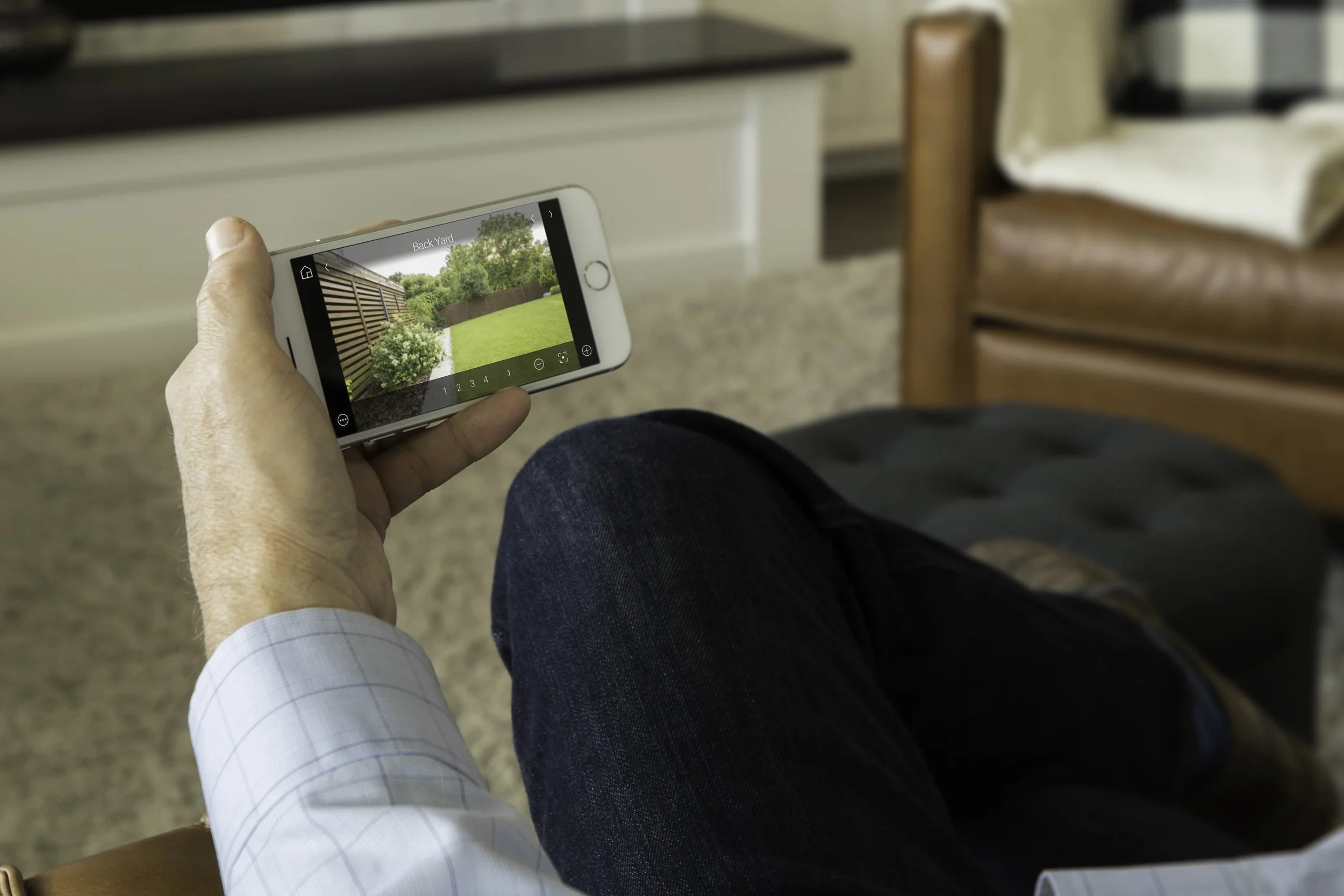 Person sitting indoors, holding a smartphone displaying a backyard with a lawn, white flowers, a wooden fence, and trees.
