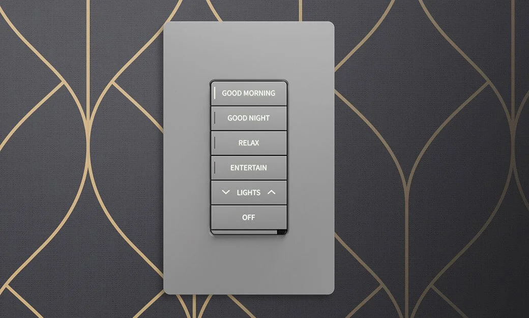 A modern wall switch panel with multiple buttons labeled 'Good Morning,' 'Good Night,' 'Relax,' 'Entertain,' 'Lights,' and 'Off,' mounted on a dark wallpaper with gold geometric patterns.
